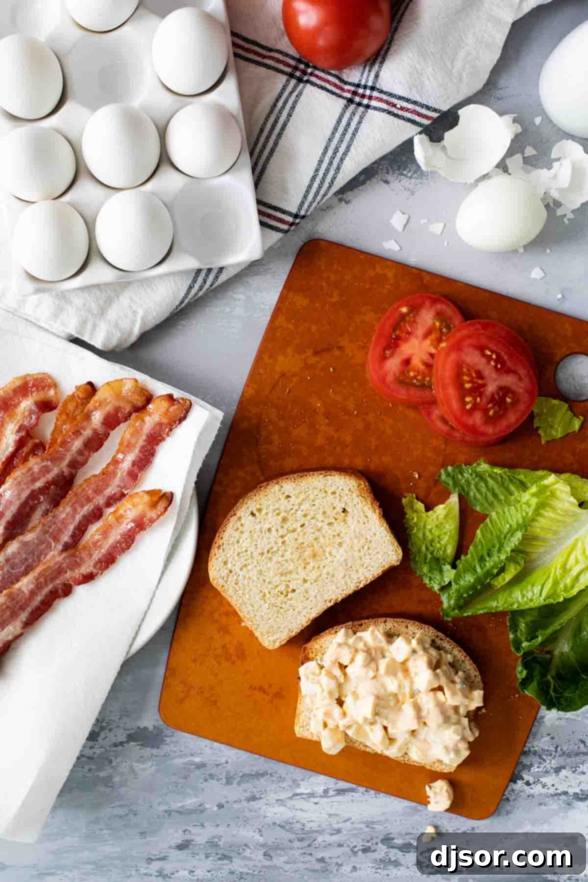 Fresh ingredients for Egg Salad BLTs arranged on a surface, including eggs, bacon, lettuce, tomato, and sauces.