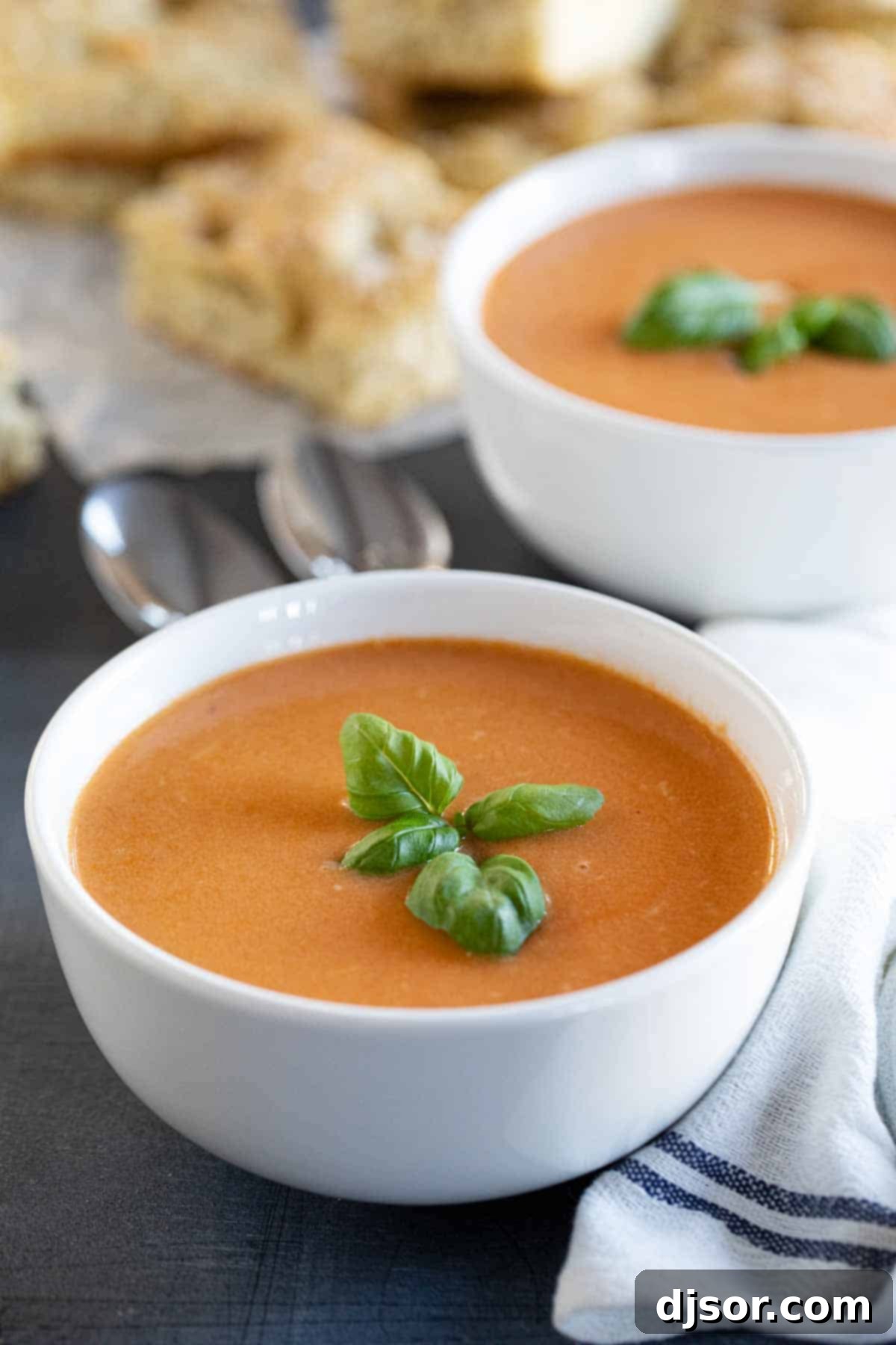 An easy slow cooker soup recipe, this Tomato Basil Soup is full of tomato flavor, is super creamy, and is easily a family favorite! Two bowls of creamy tomato basil soup garnished with fresh basil.