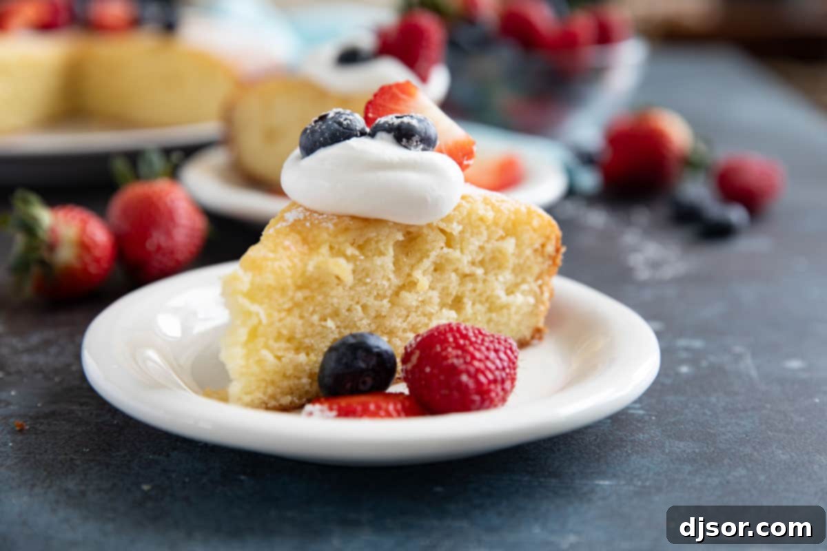 A beautifully sliced Lemon Yogurt Cake, adorned with a dusting of powdered sugar and fresh berries including blueberries, strawberries, and raspberries, topped with a dollop of whipped cream.