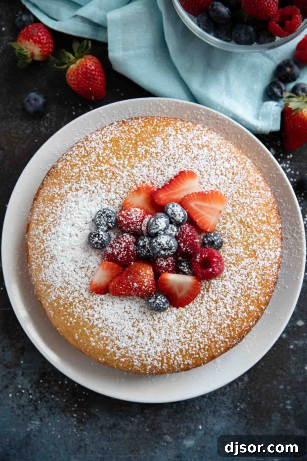 A perfectly baked and glazed Lemon Yogurt Cake resting on a white plate, elegantly garnished with a light dusting of powdered sugar and a vibrant mix of fresh berries.