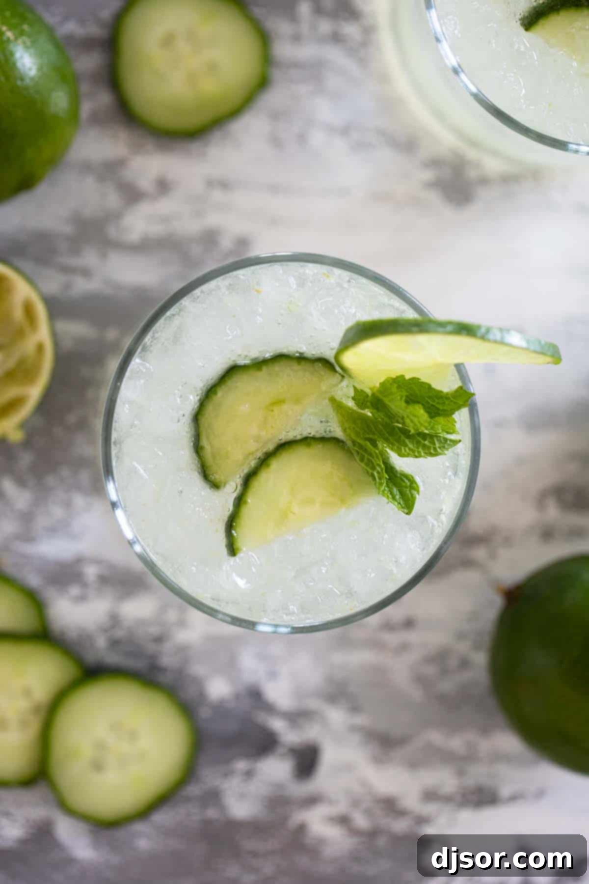 Cucumber Lime Chill 7 An overhead shot of a glass filled with cucumber limeade, ice cubes, and thin cucumber slices, garnished with a lime wedge on a dark surface.