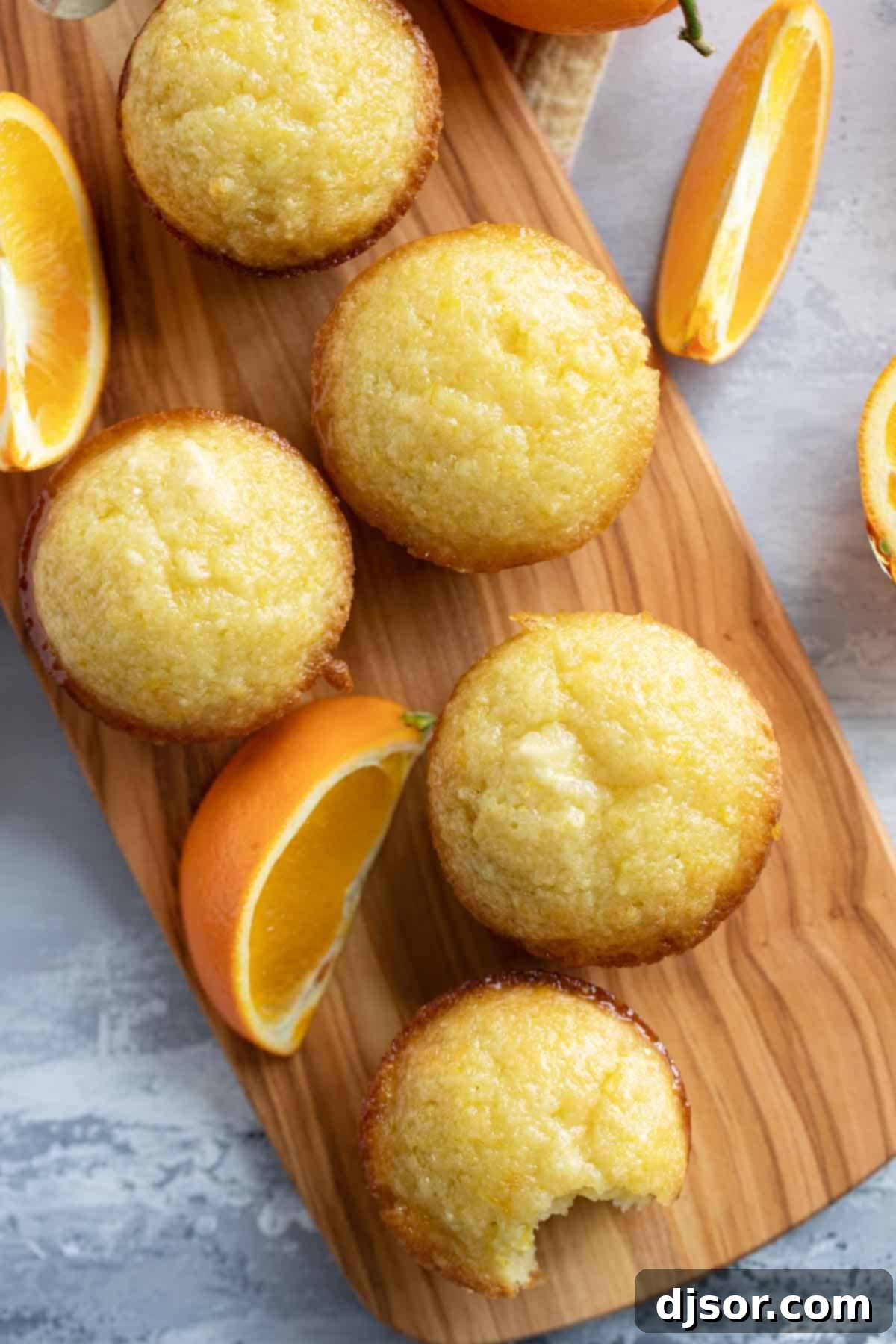 These Orange Muffins are moist and flavorful, slightly sweet with an orange simple syrup for the perfect finishing touch. A vibrant display of fresh Orange Muffins, garnished with orange slices, artfully arranged on a rustic long cutting board, ready to be enjoyed.