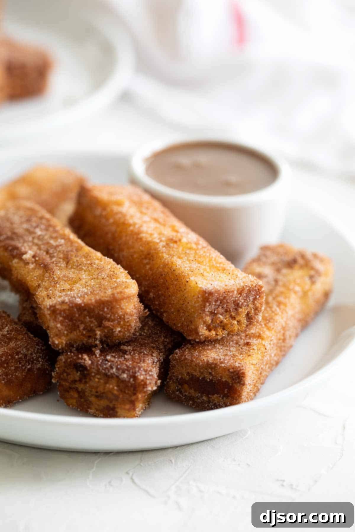 Spoil your family (or yourself!) for breakfast with these French Toast Sticks. Bread is coated in a batter and lightly fried, then covered in cinnamon sugar in this delicious breakfast treat. Golden-brown French Toast Sticks on a white plate, with a small bowl of creamy buttermilk syrup in the background for dipping.