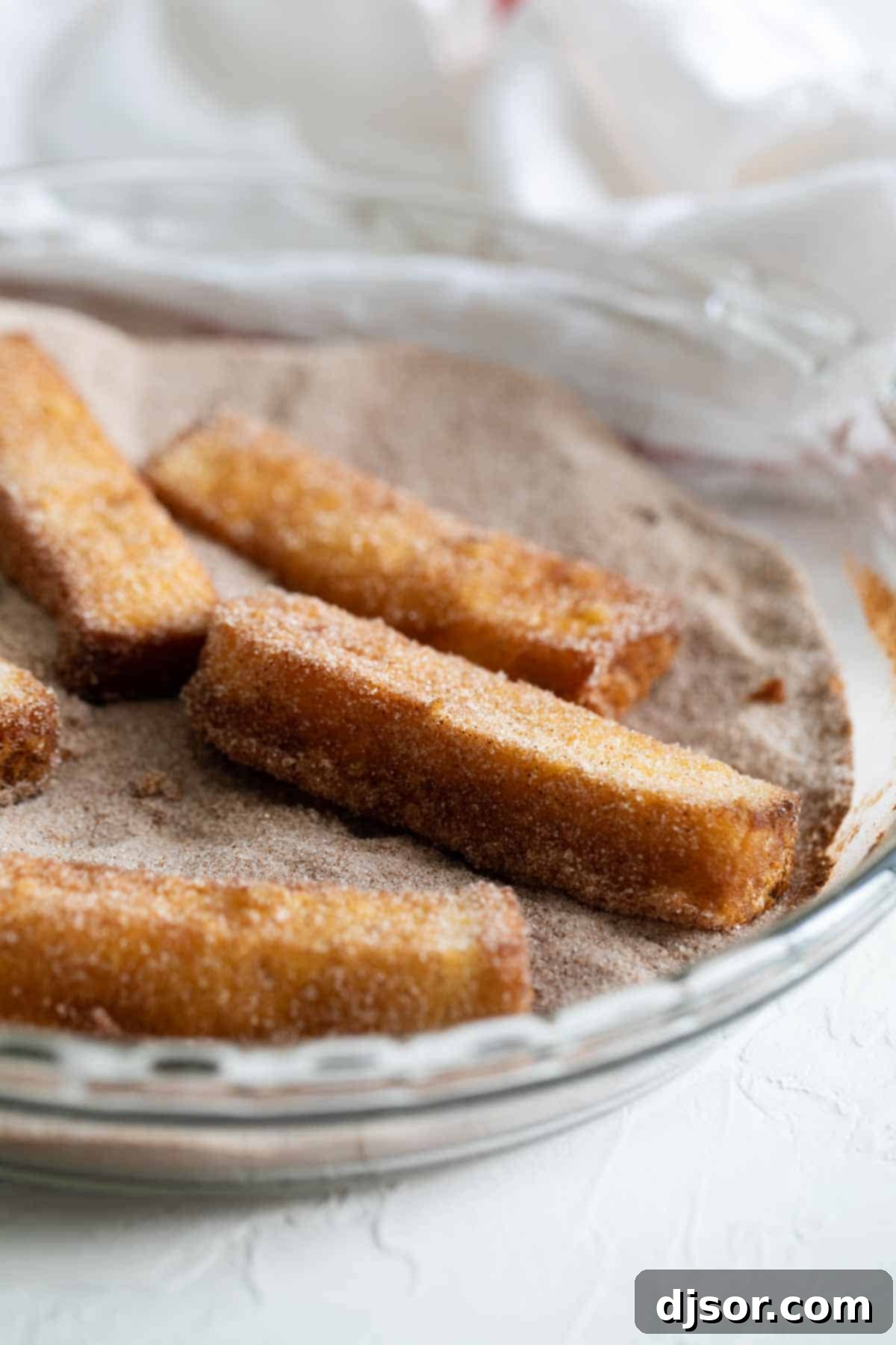 Spoil your family (or yourself!) for breakfast with these French Toast Sticks. Bread is coated in a batter and lightly fried, then covered in cinnamon sugar in this delicious breakfast treat. Close-up of French Toast Sticks being generously covered in a mixture of cinnamon sugar on a flat surface.