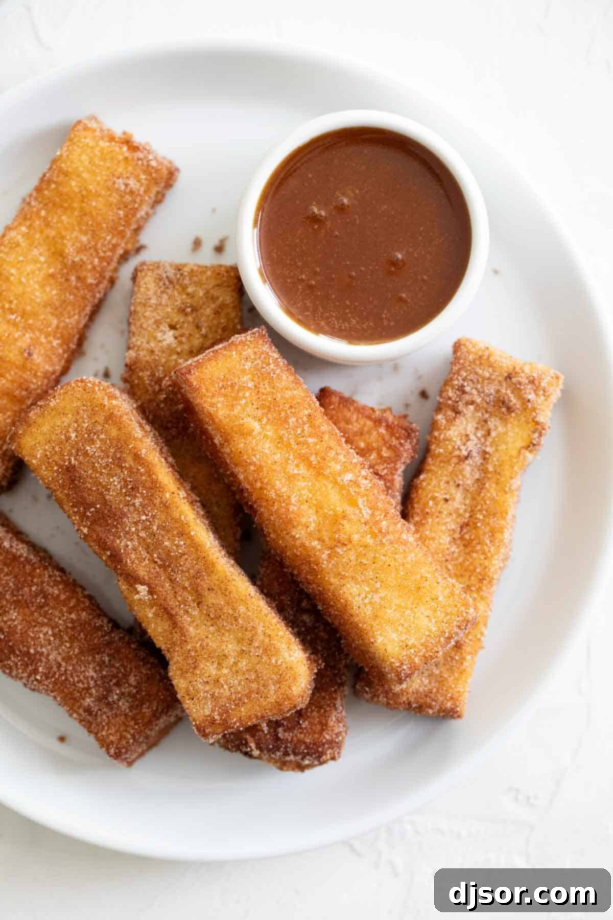 Spoil your family (or yourself!) for breakfast with these French Toast Sticks. Bread is coated in a batter and lightly fried, then covered in cinnamon sugar in this delicious breakfast treat. A stack of golden French Toast Sticks on a white plate, with a small bowl of buttermilk syrup and a fresh strawberry in the foreground.