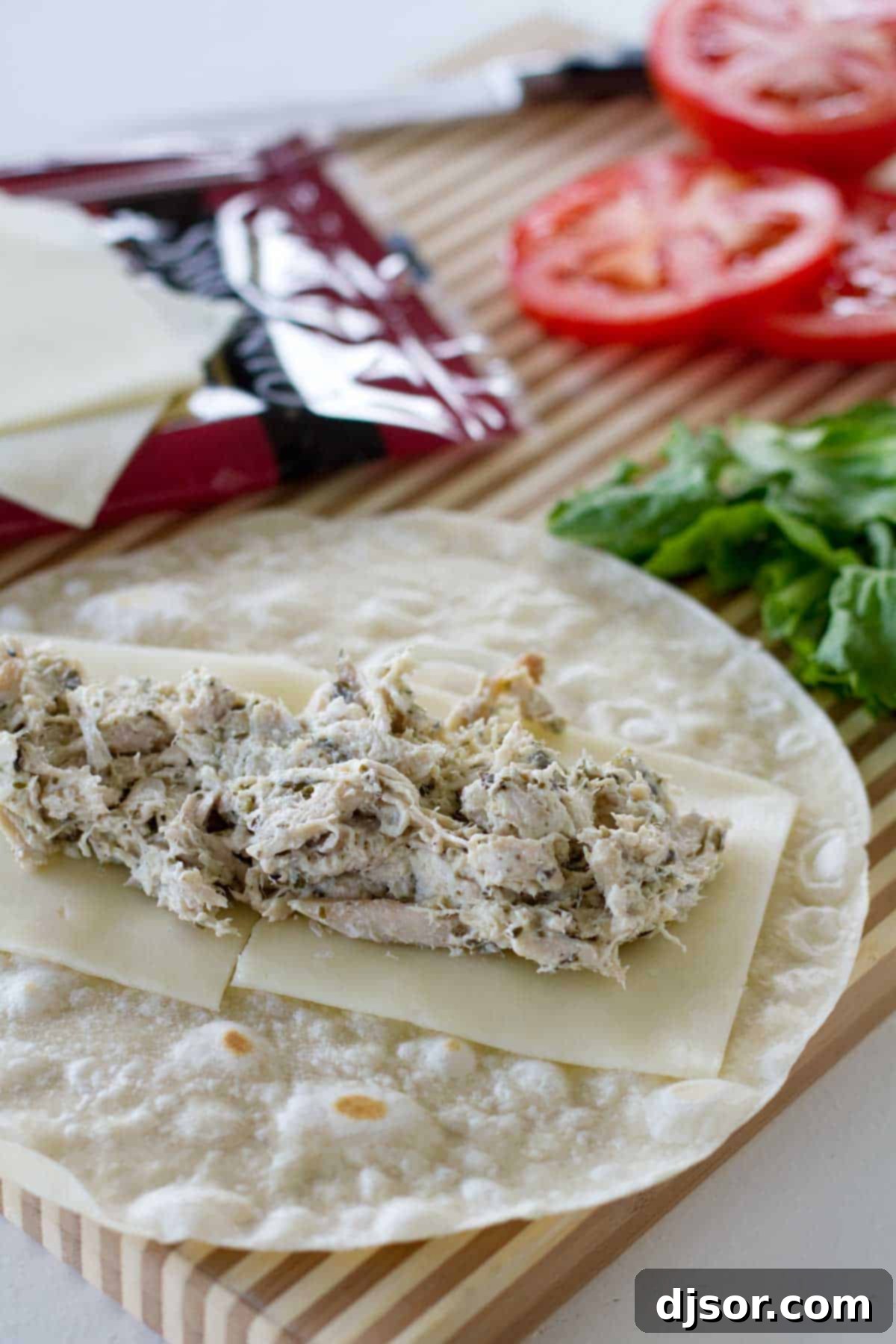 Hands assembling Chicken Pesto Wraps, layering chicken mixture, cheese, tomatoes, and lettuce on a tortilla.