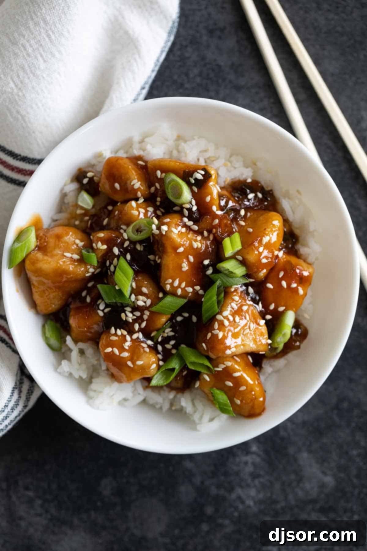 An overhead shot of a serving bowl filled with steaming white rice, topped generously with General Tso's Chicken, vibrant green onions, and toasted sesame seeds.