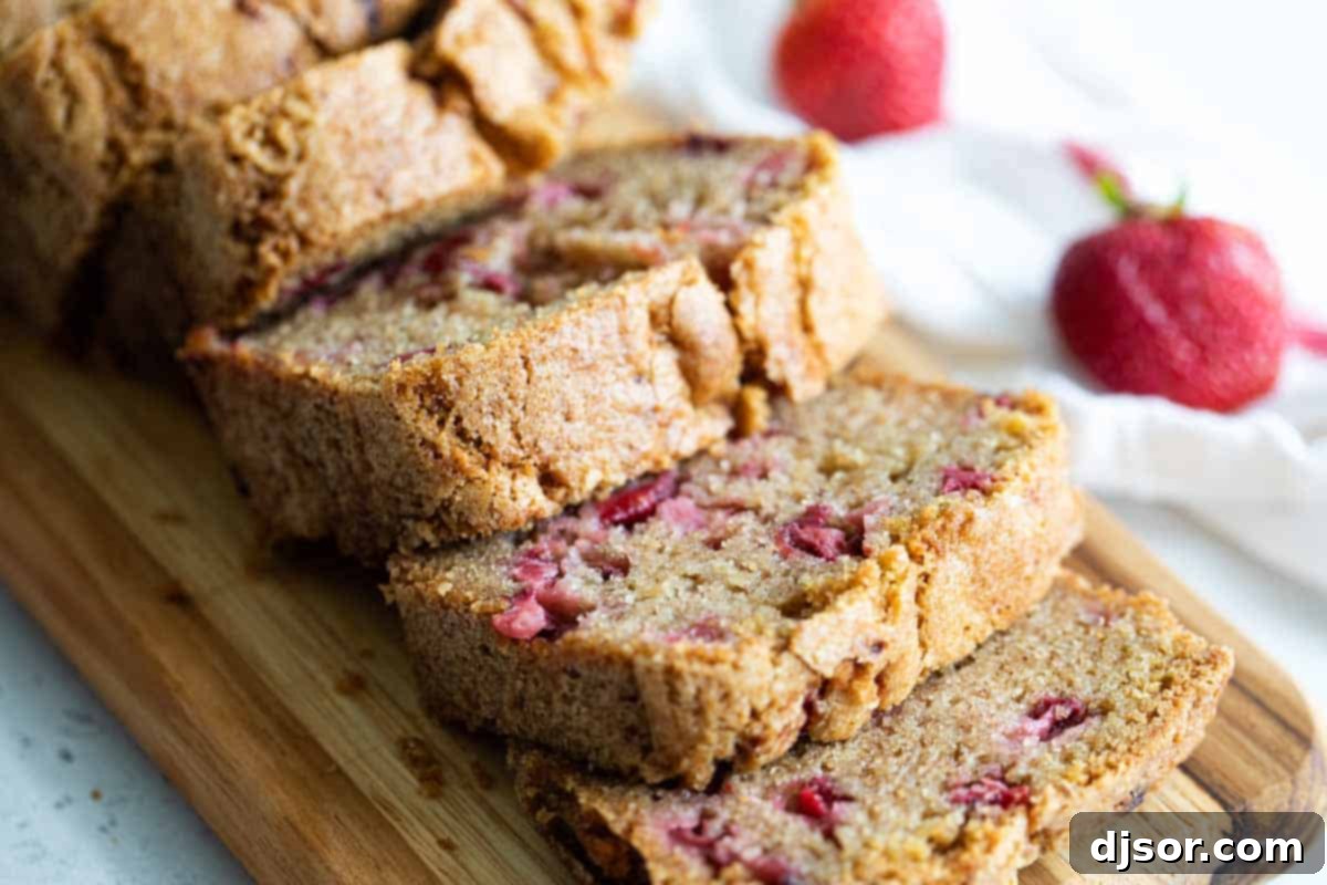 A perfectly baked loaf of strawberry bread, sliced and showing off generous chunks of fresh strawberries inside, with whole fresh strawberries scattered behind it.
