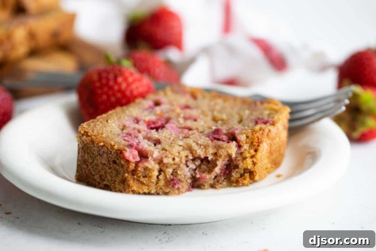 A delectable slice of strawberry bread, displaying its moist texture and visible strawberry pieces, rests on a white plate with a bite taken out of it.