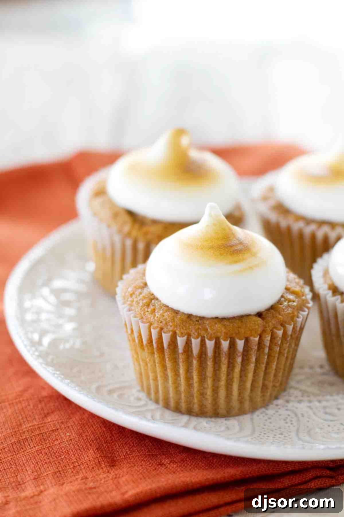 Cozy Sweet Potato Cupcakes with Torched Mallow Swirls 4 Sweet Potato Cupcakes topped with toasted marshmallow frosting, arranged neatly on a white plate, ready to be served.