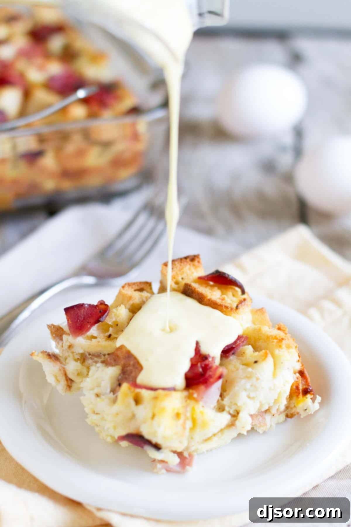 Eggs benedict made easy!! An Eggs Benedict Casserole is an overnight casserole with the flavors of eggs benedict that is served with an easy blender hollandaise sauce poured over the top. A close-up shot of rich, golden hollandaise sauce being poured over a warm slice of Eggs Benedict Casserole, creating an enticing cascade.