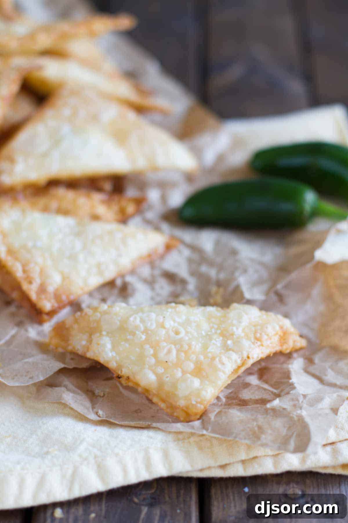 These Jalapeno Popper Wontons are fried wontons that are stuffed with cream cheese and jalapeños in this tasty appetizer. Jalapeno Popper Wontons resting on brown paper, sprinkled with coarse salt.