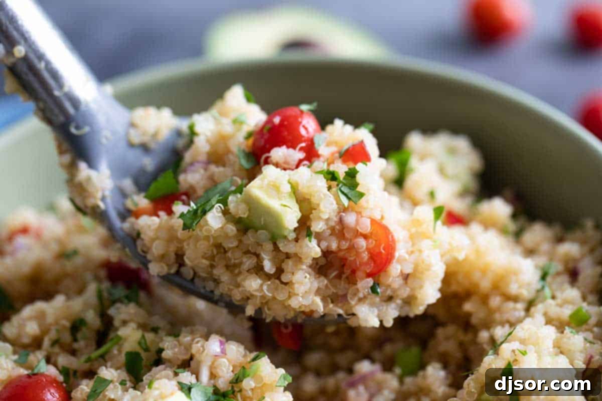 A serving spoon lifting a scoop of quinoa salad, showing avocado, tomato, and bell pepper.