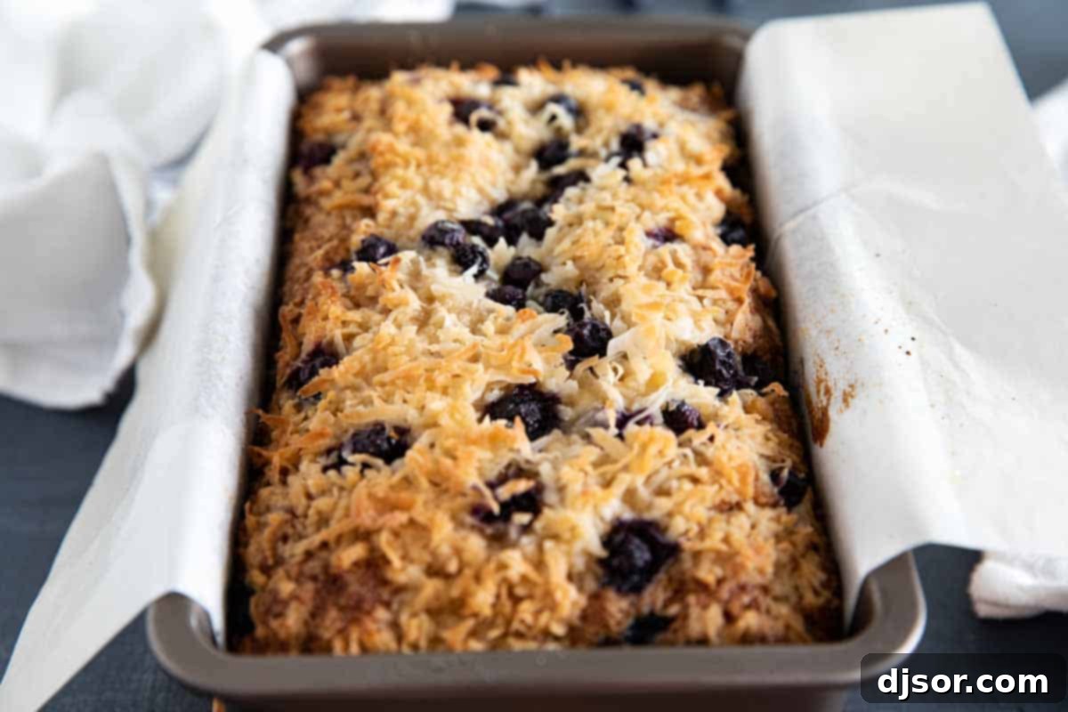 Make a change from your typical banana bread with this Blueberry Banana Bread with Coconut that is filled with fresh berries and topped with a crunchy streusel topping. A freshly baked loaf of blueberry banana bread with coconut, still in its parchment-lined baking pan, cooling on a wire rack, with its golden-brown streusel topping visible.