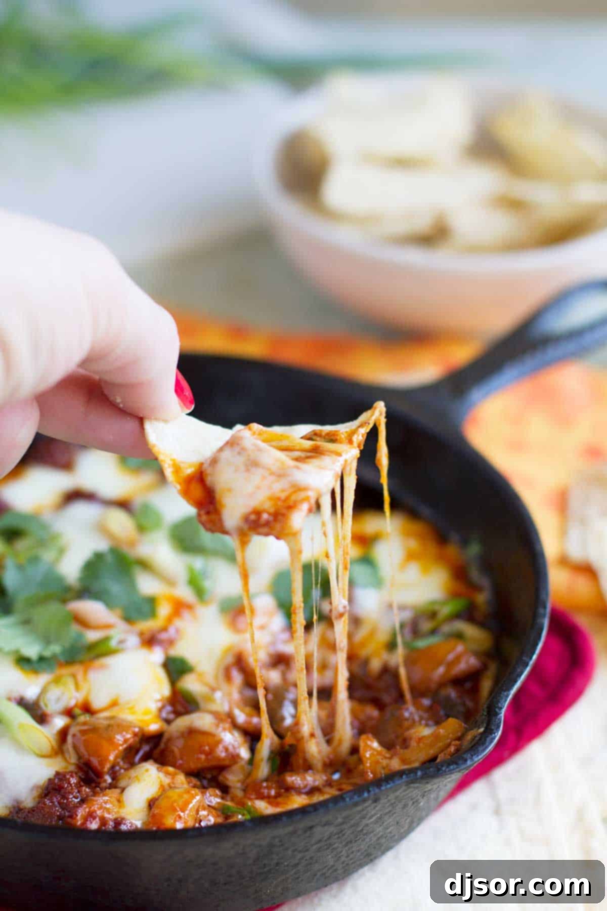 A hand holding a crispy tortilla chip, perfectly dipped into the warm, gooey Mushroom and Chorizo Queso Dip, showing its rich, cheesy texture.