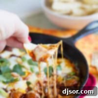 A hand with a tortilla chip dipping into a bubbly Mushroom and Chorizo Queso Dip, garnished with green onions and cilantro.