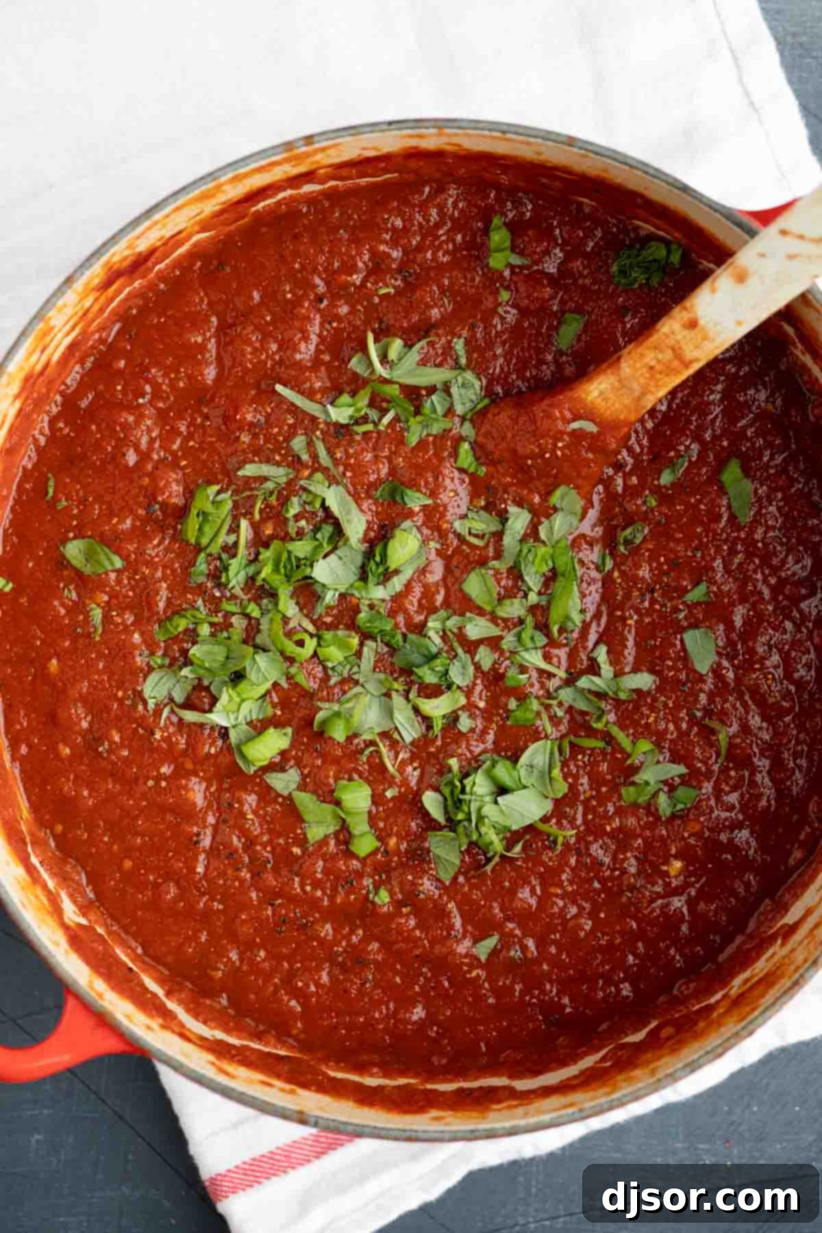 Overhead view of a rustic Dutch oven brimming with freshly made, vibrant red marinara sauce, garnished with fresh basil leaves.