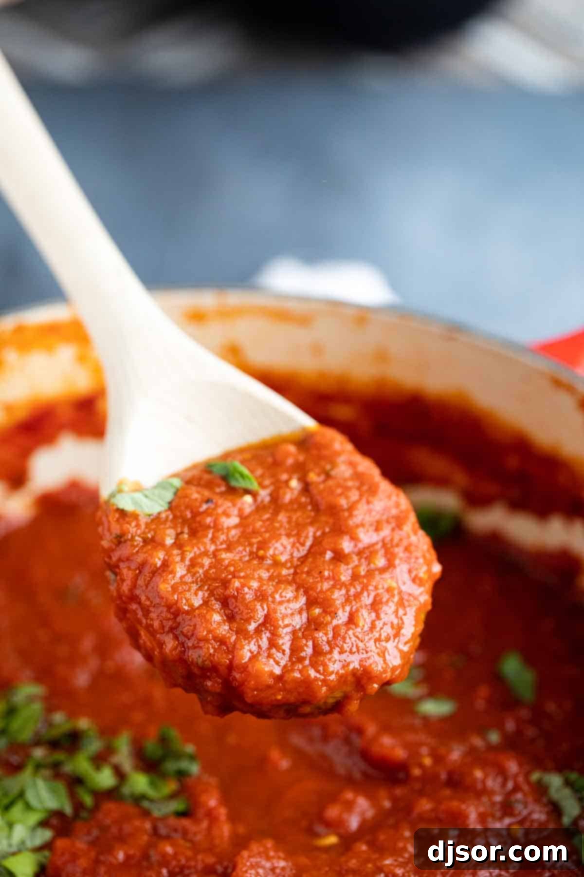 A wooden spoon lifted from a pot, showcasing a generous scoop of homemade marinara sauce, garnished with a sprinkle of fresh basil.