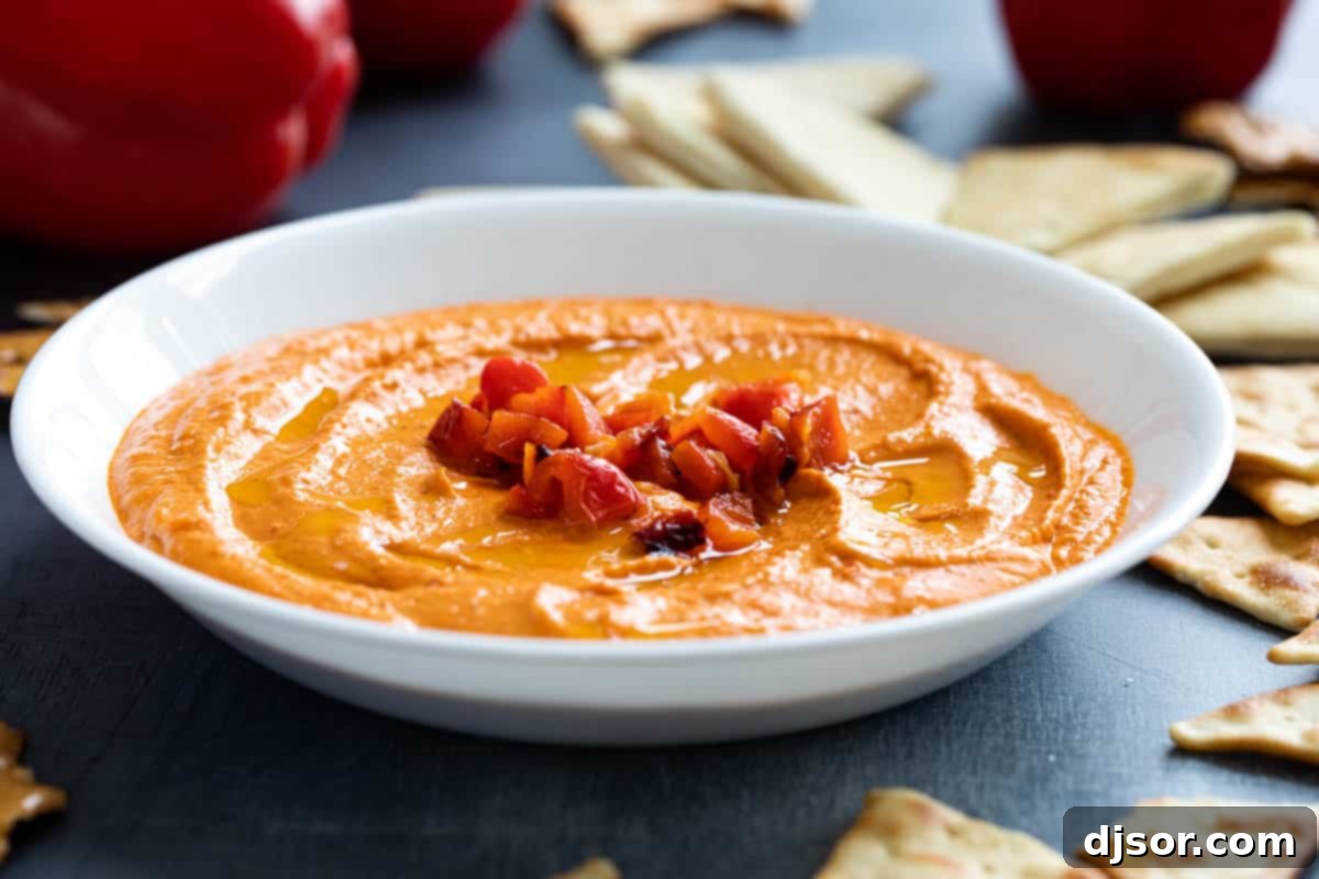 Whether you are hosting gathering or craving a healthy snack, this Roasted Red Pepper Hummus is what you need! Smoky roasted red peppers are combined with creamy chickpeas and spices to make this dip that is perfect for crackers, bread, or veggies. Homemade roasted red pepper hummus topped with chunks of roasted red peppers in a rustic bowl.