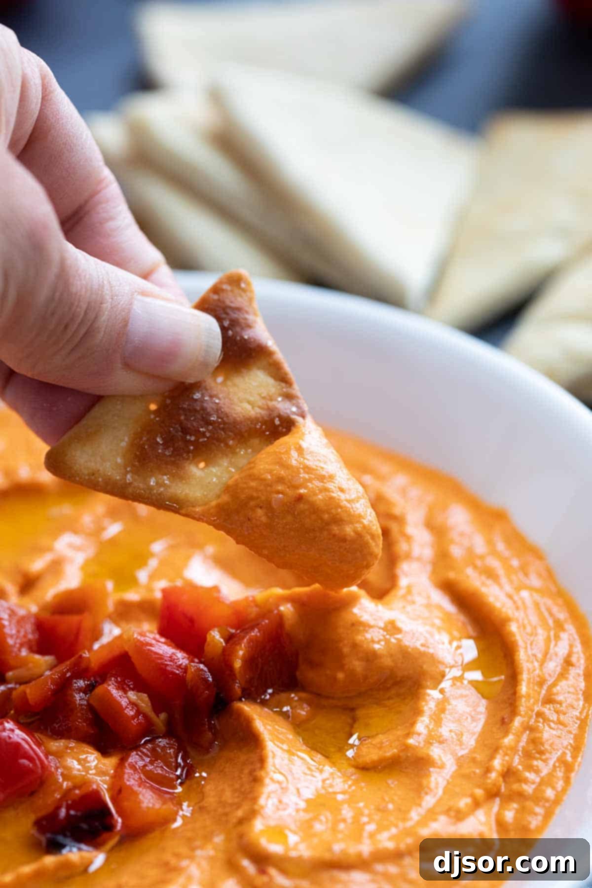 Dipping a pita chip into roasted red pepper hummus. A hand dipping a crispy pita chip into a bowl of creamy roasted red pepper hummus.