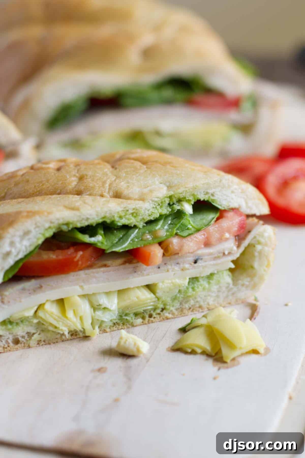 French bread is stuffed with artichoke hearts, turkey and vegetables and then slathered with an easy basil sauce in this easy Turkey Sub with Artichokes and Basil that’s perfect for a picnic or for dinner. A vibrant turkey sub, generously filled with artichoke hearts, turkey, and fresh vegetables, is artfully sliced into individual serving portions on a wooden cutting board, showcasing its rich layers and appealing presentation.