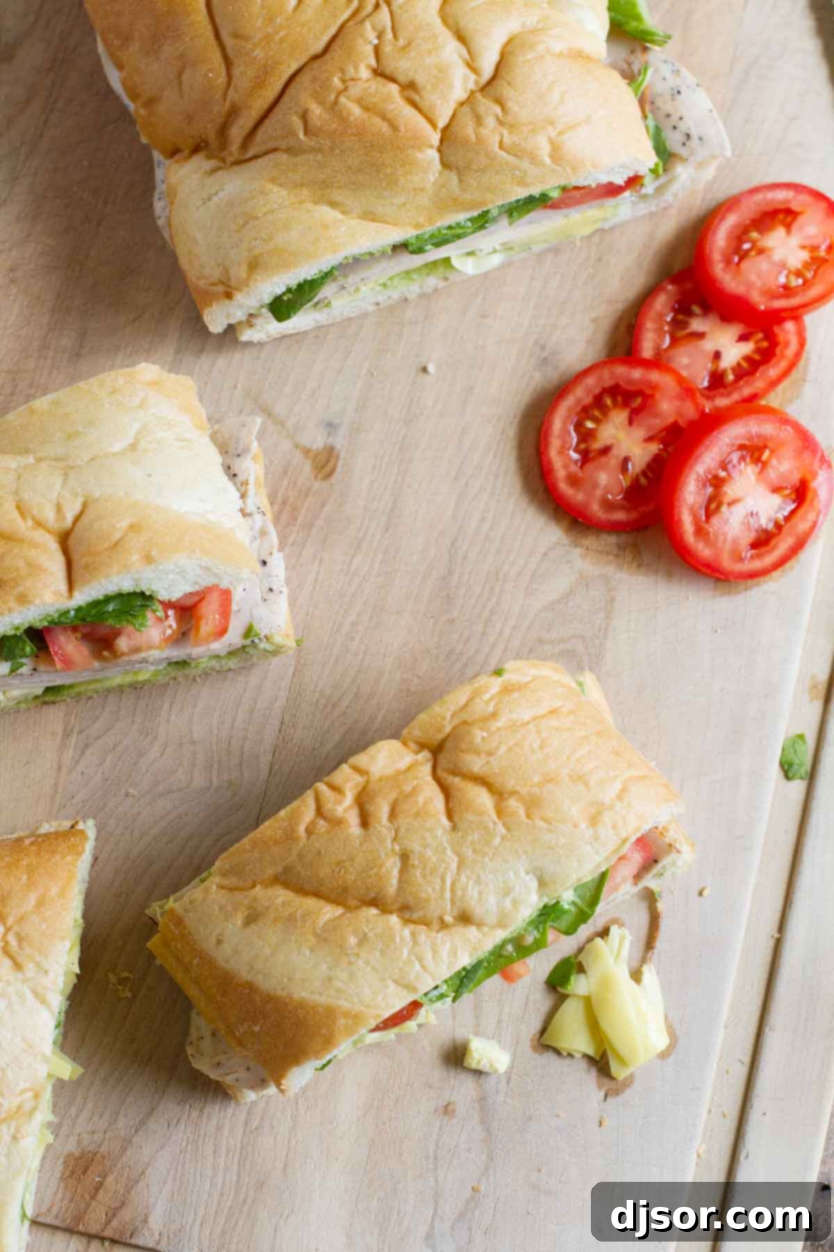 French bread is stuffed with artichoke hearts, turkey and vegetables and then slathered with an easy basil sauce in this easy Turkey Sub with Artichokes and Basil that’s perfect for a picnic or for dinner. A large, gourmet turkey sub, generously stuffed with artichoke hearts, slices of deli turkey, fresh tomatoes, Havarti cheese, and baby spinach, is presented on a wooden cutting board. The sub has been expertly sliced into multiple individual portions, revealing its vibrant layers and making it ready to serve as a delicious and convenient meal.
