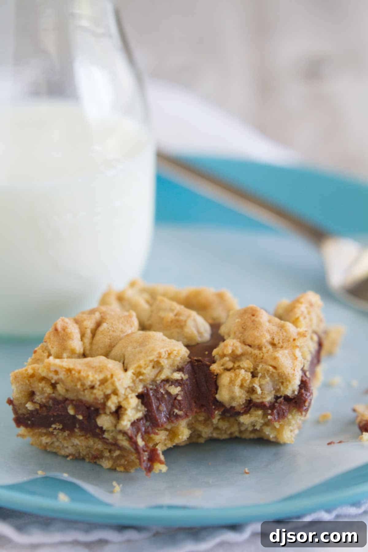 Gooey chocolate is sandwiched between two layers of oatmeal cookies in these decadent Oatmeal Fudge Bars. A single Oatmeal Fudge Bar on a blue plate with a bite taken from it, showing the layers.