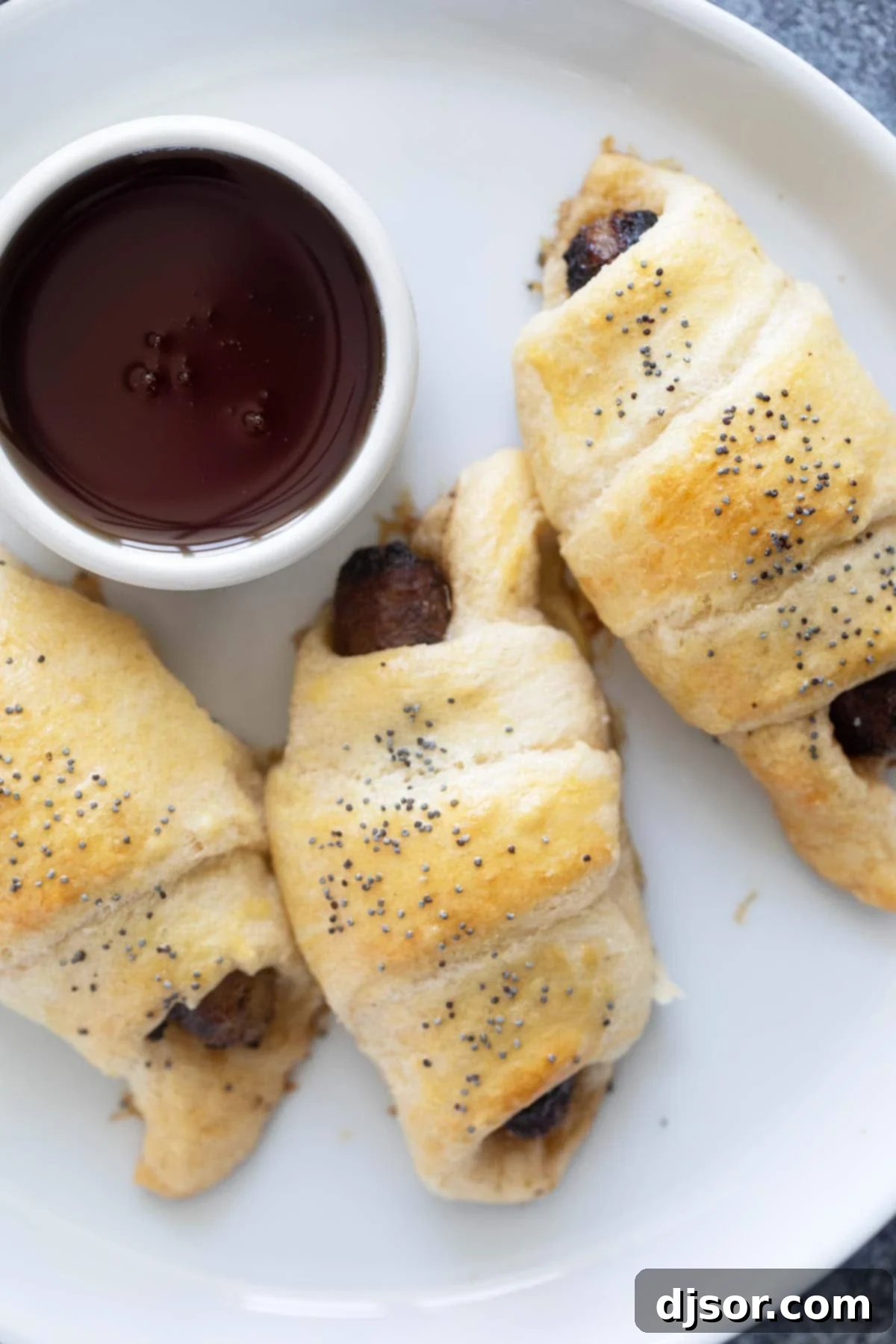 Fluffy Breakfast Sausage Rolls 6 A single Breakfast Pig in a Blanket on a white plate, ready to be picked up, with a small ramekin of maple syrup for dipping.