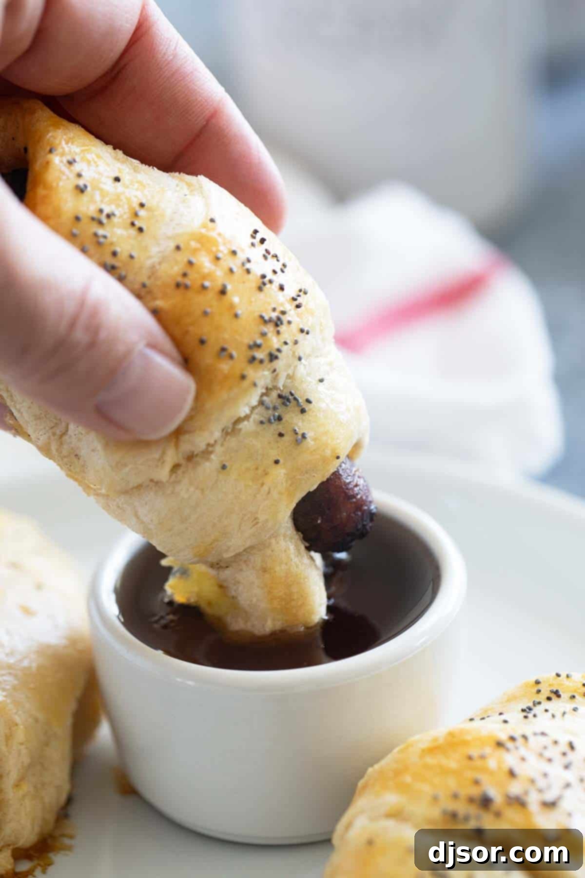 Fluffy Breakfast Sausage Rolls 7 A close-up shot of a hand dipping a golden brown Breakfast Pig in a Blanket into a small bowl of rich maple syrup.