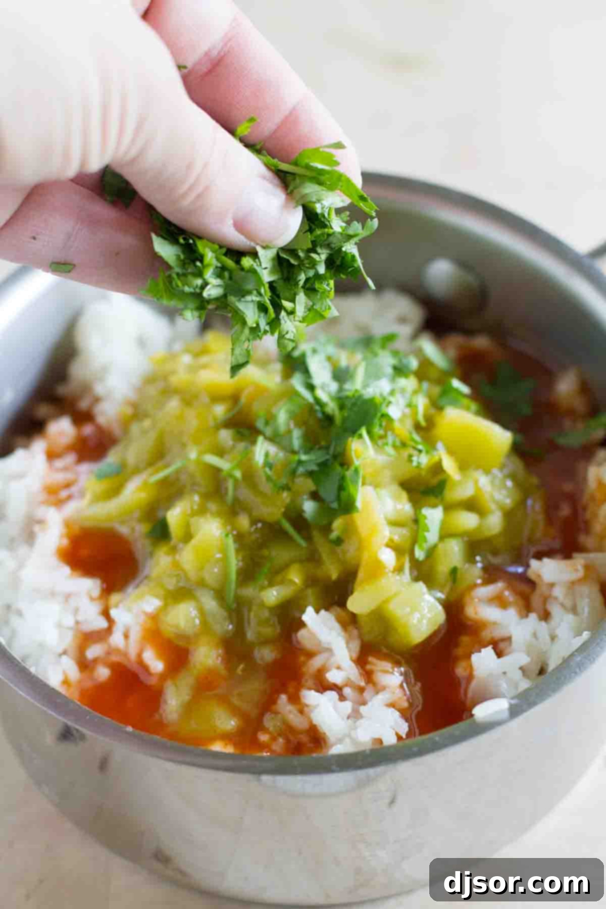 A close-up view of a pot of fluffy cooked rice, into which vibrant red enchilada sauce, diced green chiles, and fresh chopped cilantro are being stirred, illustrating the final mixing step of the Enchilada Rice recipe.