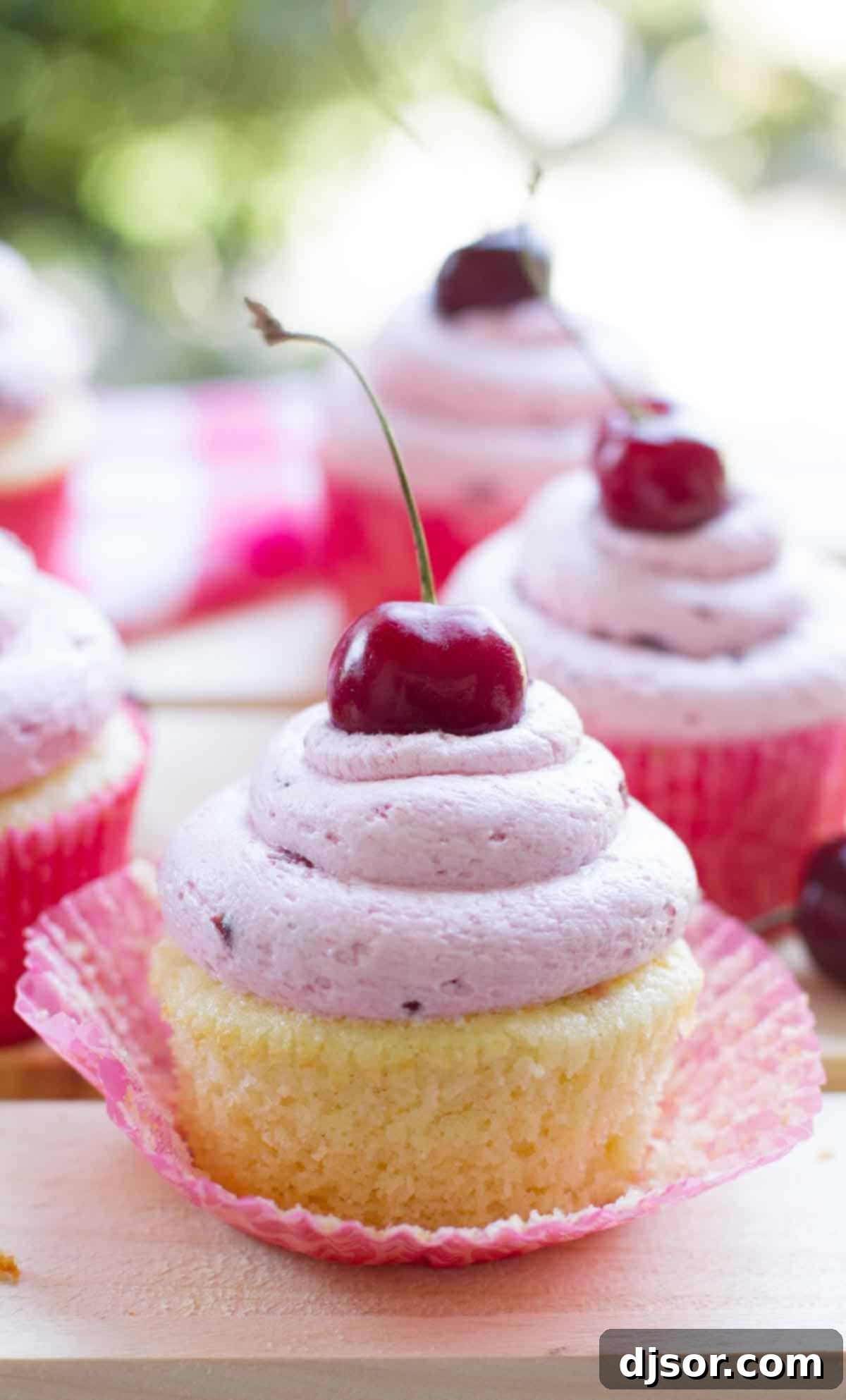Close-up of a moist almond cupcake with its paper liner pulled back, revealing a delicate crumb, topped with fresh cherry frosting.