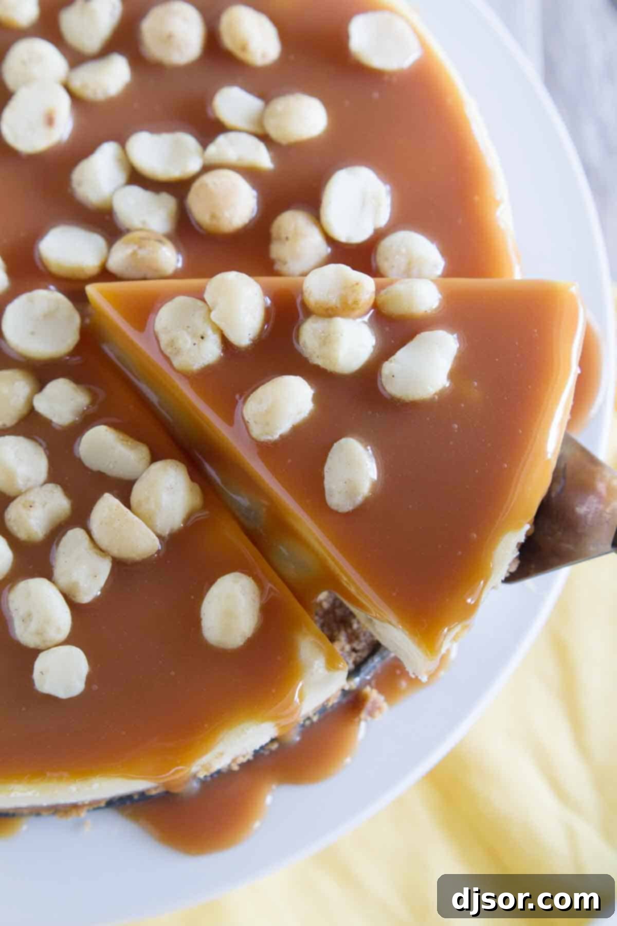 White Chocolate Cheesecake with Macadamia Nuts and Caramel - Creamy white chocolate cheesecake has macadamia nuts throughout, with a homemade caramel sauce poured over the top. A hand gently lifts a perfect slice of white chocolate macadamia nut cheesecake from the whole, revealing its creamy texture and crust.