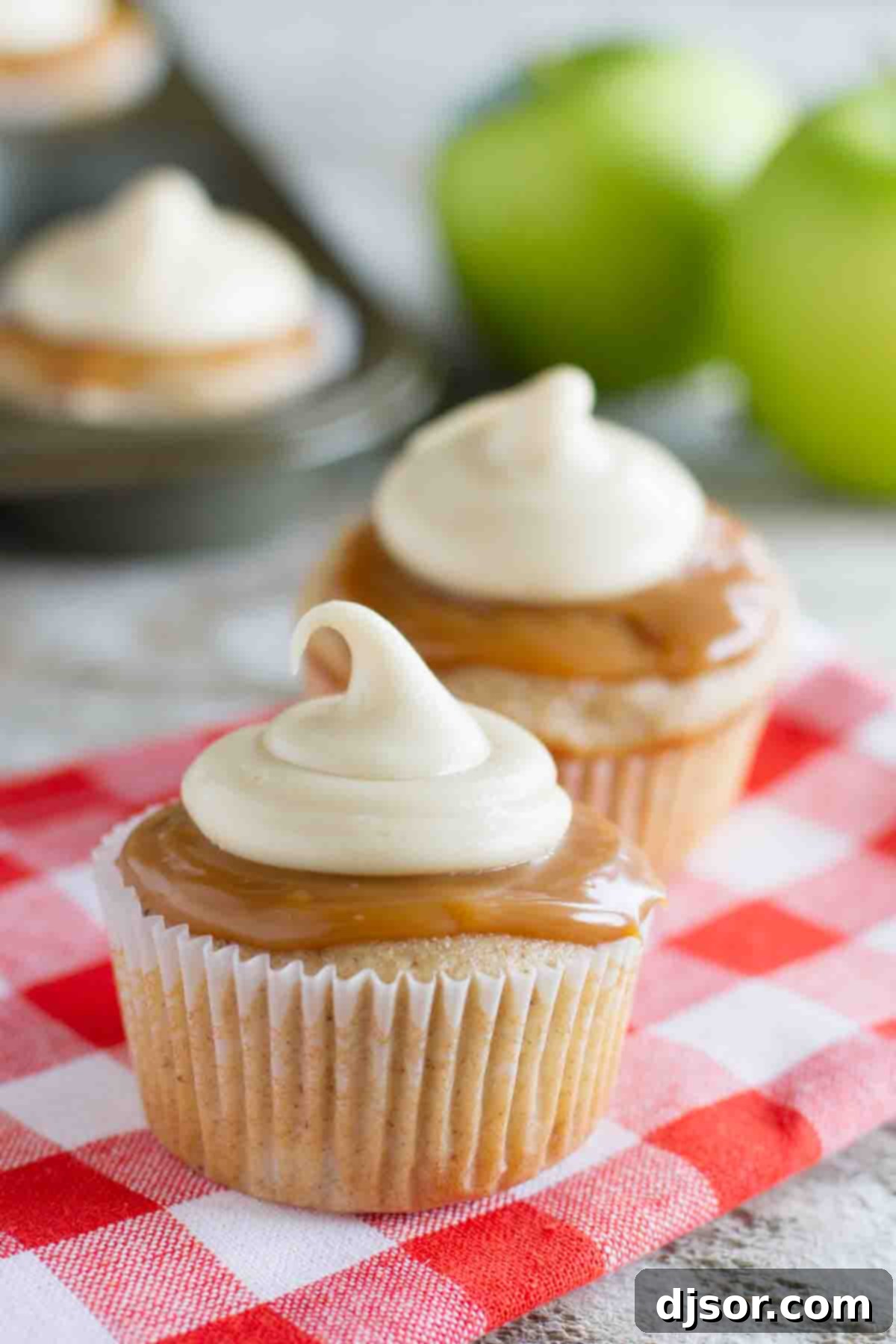 Decadent Caramel Apple Cupcakes 2 Close-up of frosted Caramel Apple Cupcakes with fresh apples and more cupcakes blurred in the background, showcasing rich caramel icing and a warm fall aesthetic.