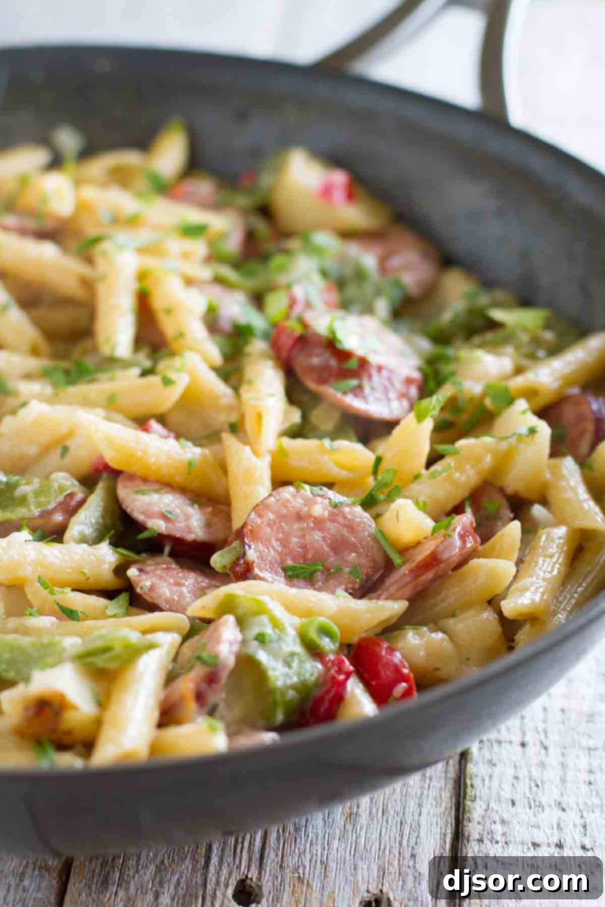 A large skillet brimming with creamy pasta and perfectly sliced smoked sausage, garnished with fresh green parsley.