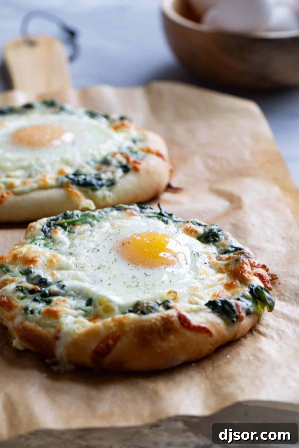 Breakfast meets dinner with this Egg Pizza with Creamed Spinach which tops pizza dough with creamed spinach and a perfectly cooked egg. Two golden-brown individual egg pizzas with creamed spinach and melted cheese on parchment paper, fresh out of the oven.