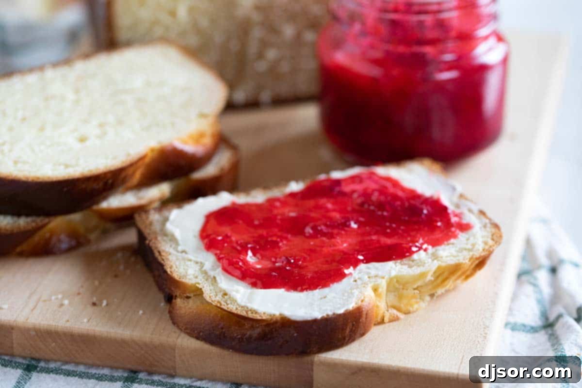 A delectable slice of golden brioche bread generously spread with butter and vibrant strawberry jam, presented on a simple white plate, ready to be enjoyed.