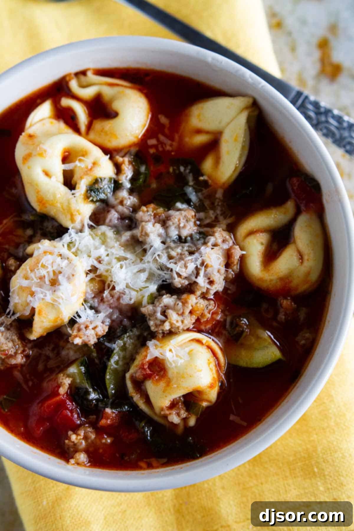A close-up shot of a bowl of Sausage Tortellini Soup, generously topped with freshly grated Parmesan cheese and a sprinkle of fresh herbs.
