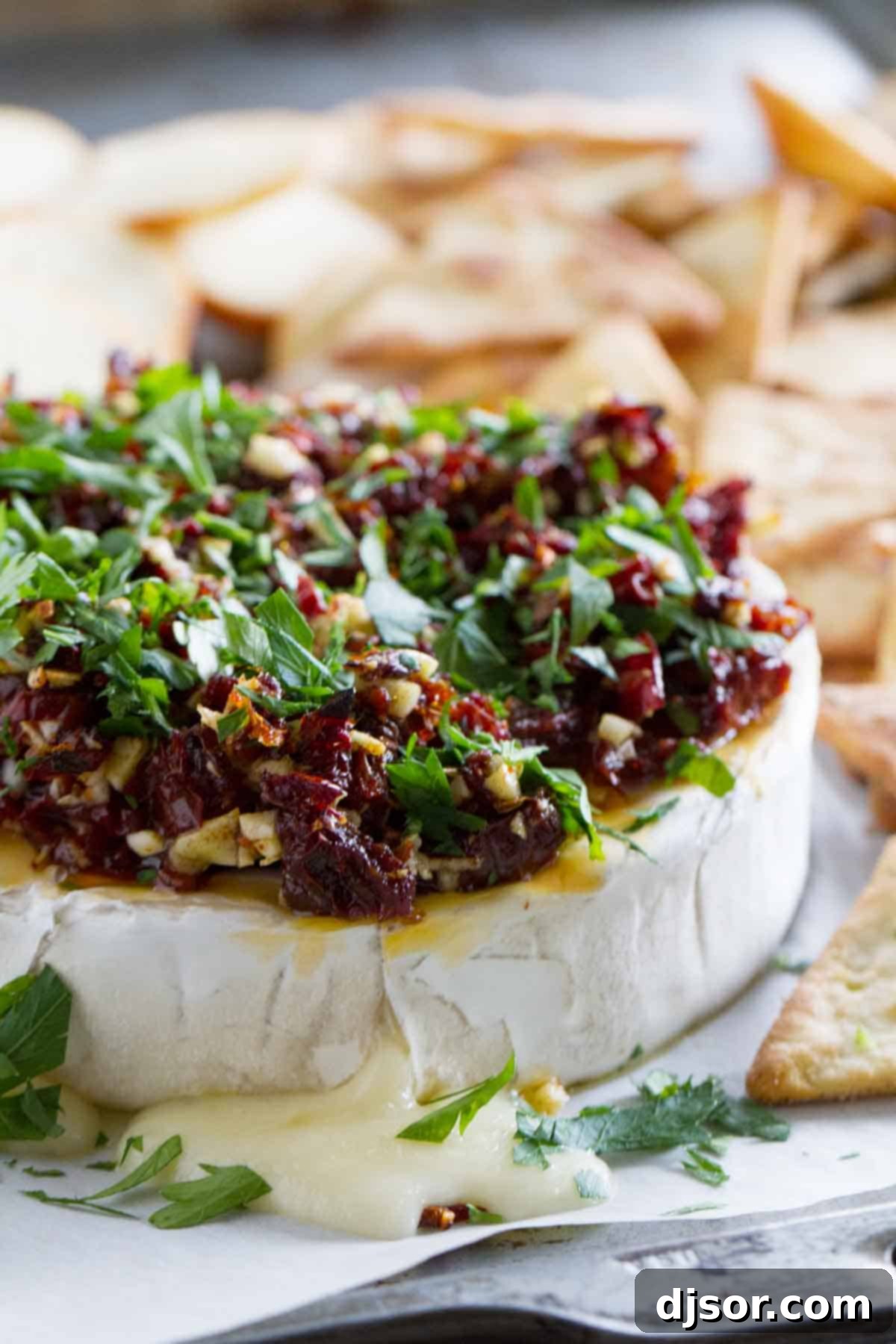 Irresistibly gooey baked brie with savory toppings. A close-up of a baked brie wheel topped with sun-dried tomatoes and herbs.