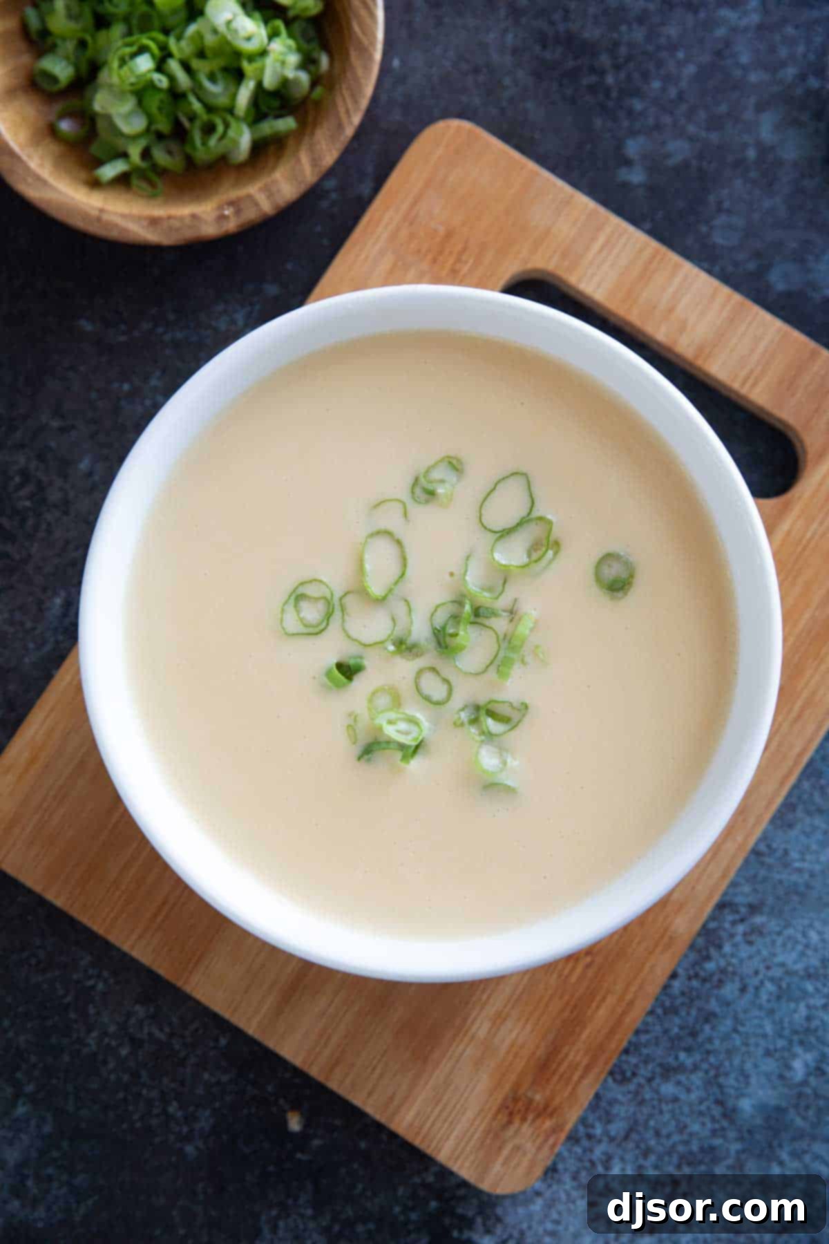 A rustic wooden cutting board holding a white bowl of golden Cheddar Cheese Soup, perfectly smooth and garnished with green onions, highlighting its rich color and inviting texture.