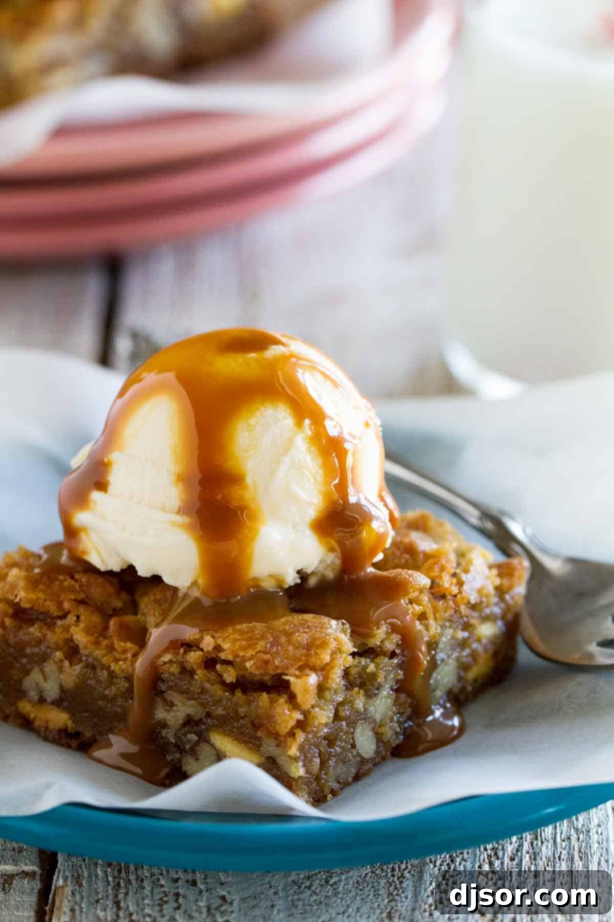 The Best Blondie Recipe with Ice Cream and Caramel A delicious blondie bar topped with a scoop of vanilla ice cream and drizzled with caramel sauce, sitting on a white plate.