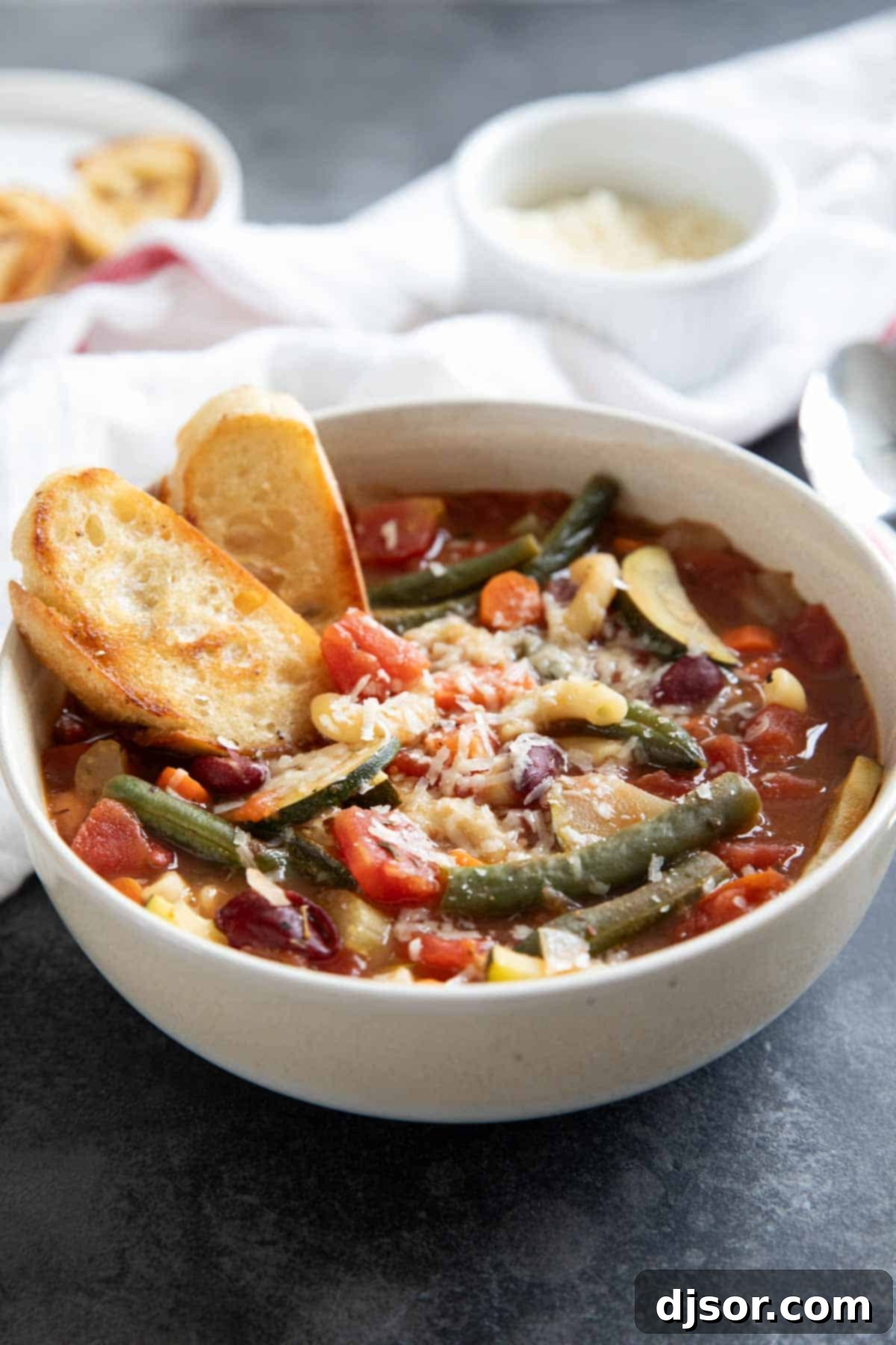 A beautifully presented bowl of Minestrone Soup, garnished with Parmesan cheese and fresh herbs, served alongside a slice of golden toasted bread for dipping.