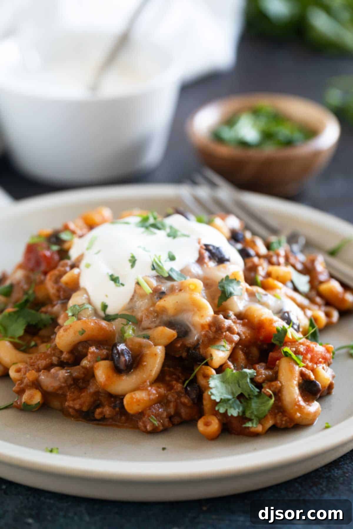 A single serving of Chili Mac on a white plate, topped with a generous dollop of sour cream and a sprig of cilantro, ready to eat.