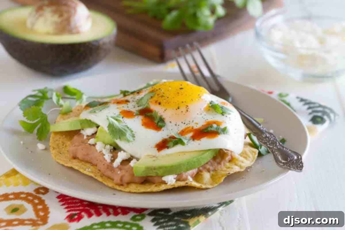 A beautifully plated breakfast tostada featuring crisp tostada shells, creamy refried beans, vibrant avocado slices, and a perfectly cooked sunny-side-up egg, garnished with fresh cilantro and a drizzle of hot sauce.