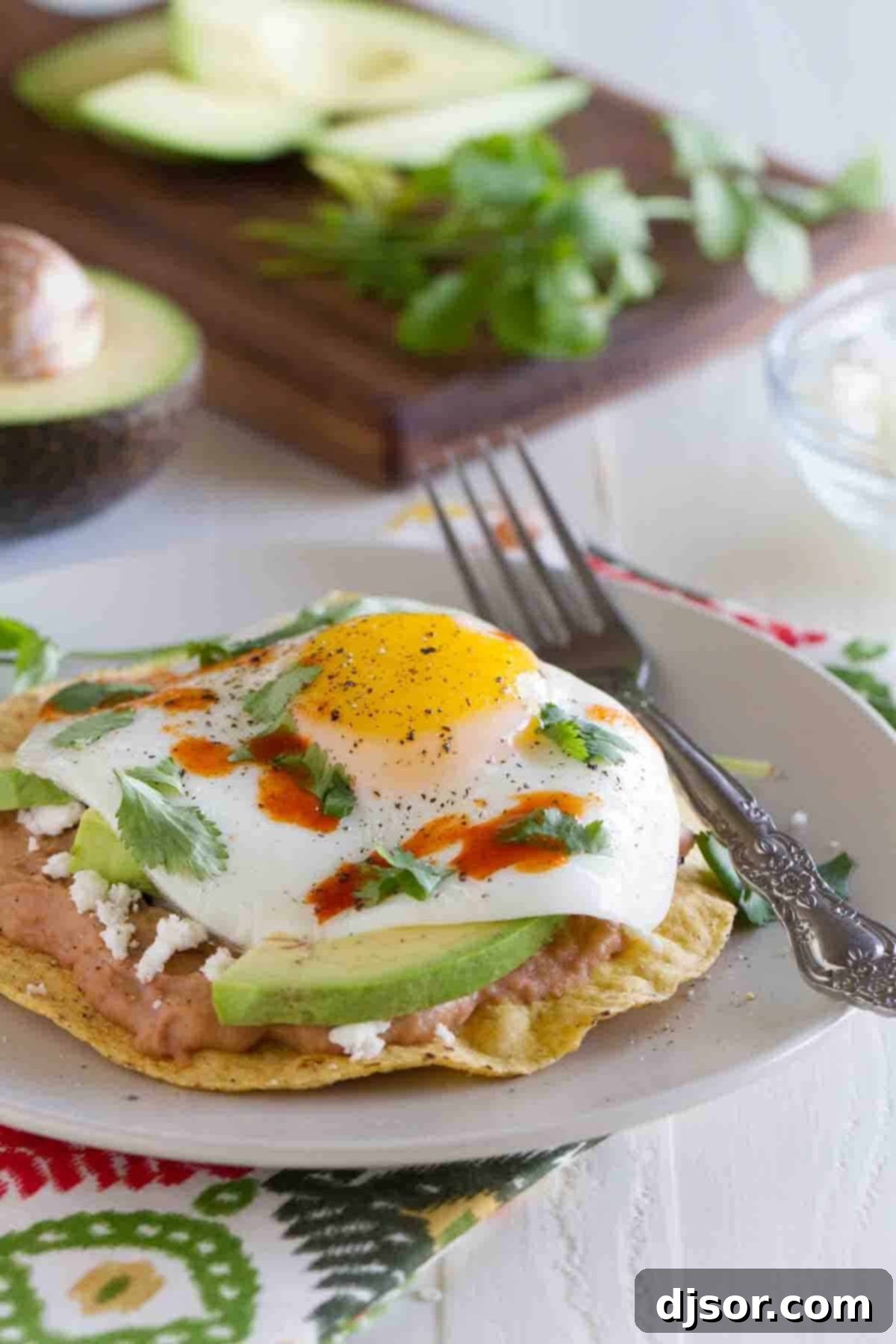 A vibrant Breakfast Tostada served on a simple white plate with a fork, ready to be enjoyed. The perfectly cooked egg yolk is visible, adding rich creaminess to the refried beans, avocado, and crispy shell.