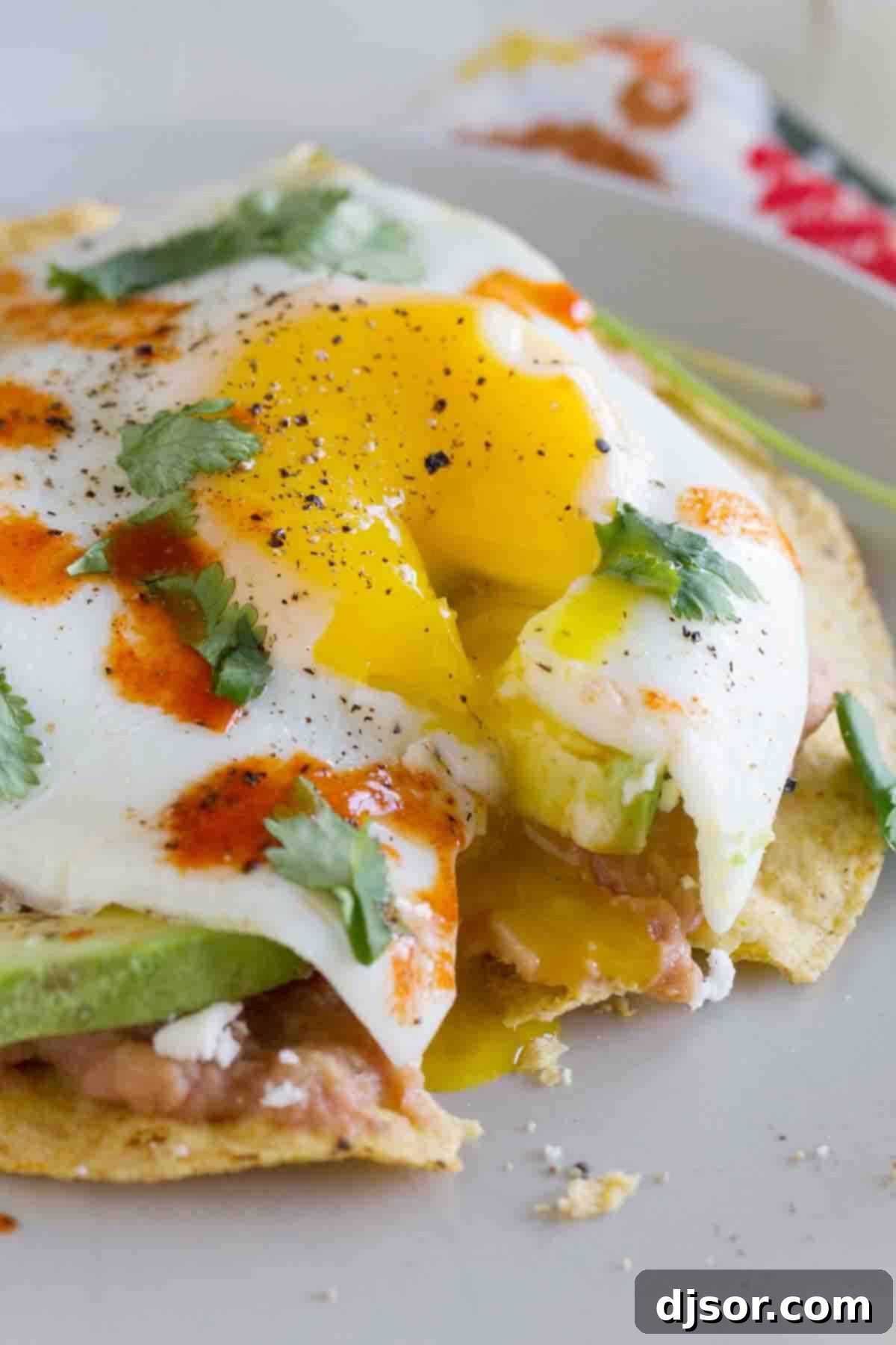 A close-up shot of a breakfast tostada with a bite taken out, showing the rich, runny yolk of the sunny-side-up egg drizzling down over the creamy refried beans and avocado, highlighting its delicious texture.