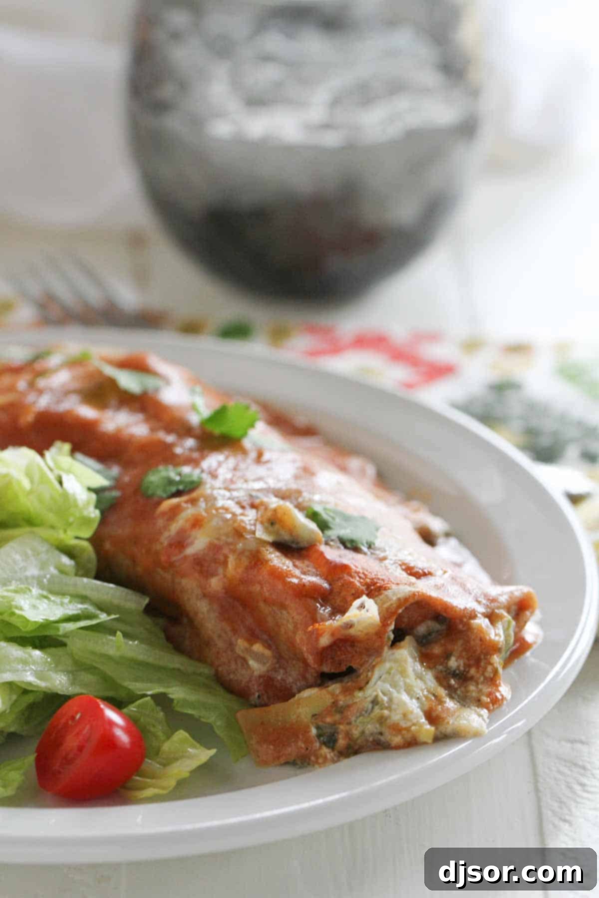 A single serving of Spinach Artichoke Enchilada presented beautifully on a white plate, accompanied by a fresh green side salad. The enchilada is generously topped with a rich sauce and melted cheese.