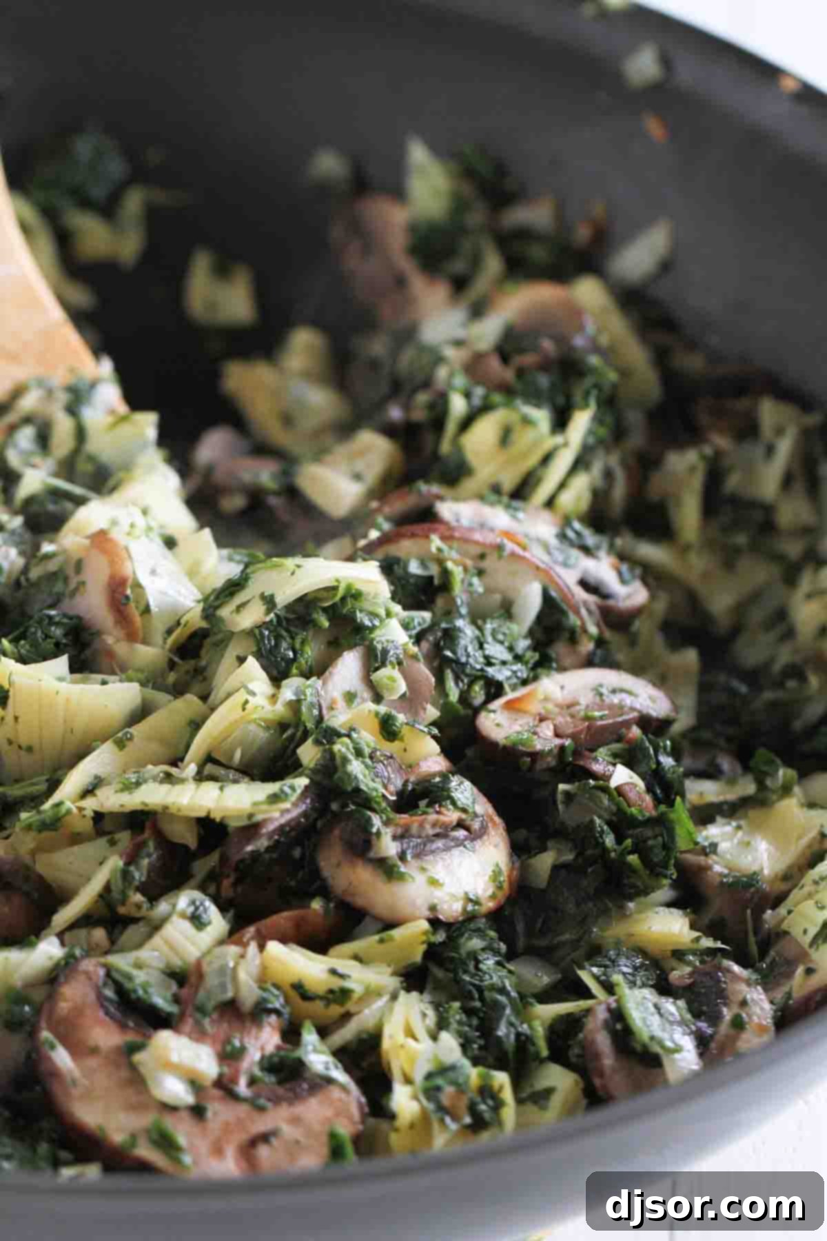 Close-up shot of the creamy and vibrant filling for spinach artichoke enchiladas being prepared in a large skillet, showcasing a mix of cooked spinach, artichoke hearts, mushrooms, onions, and garlic.