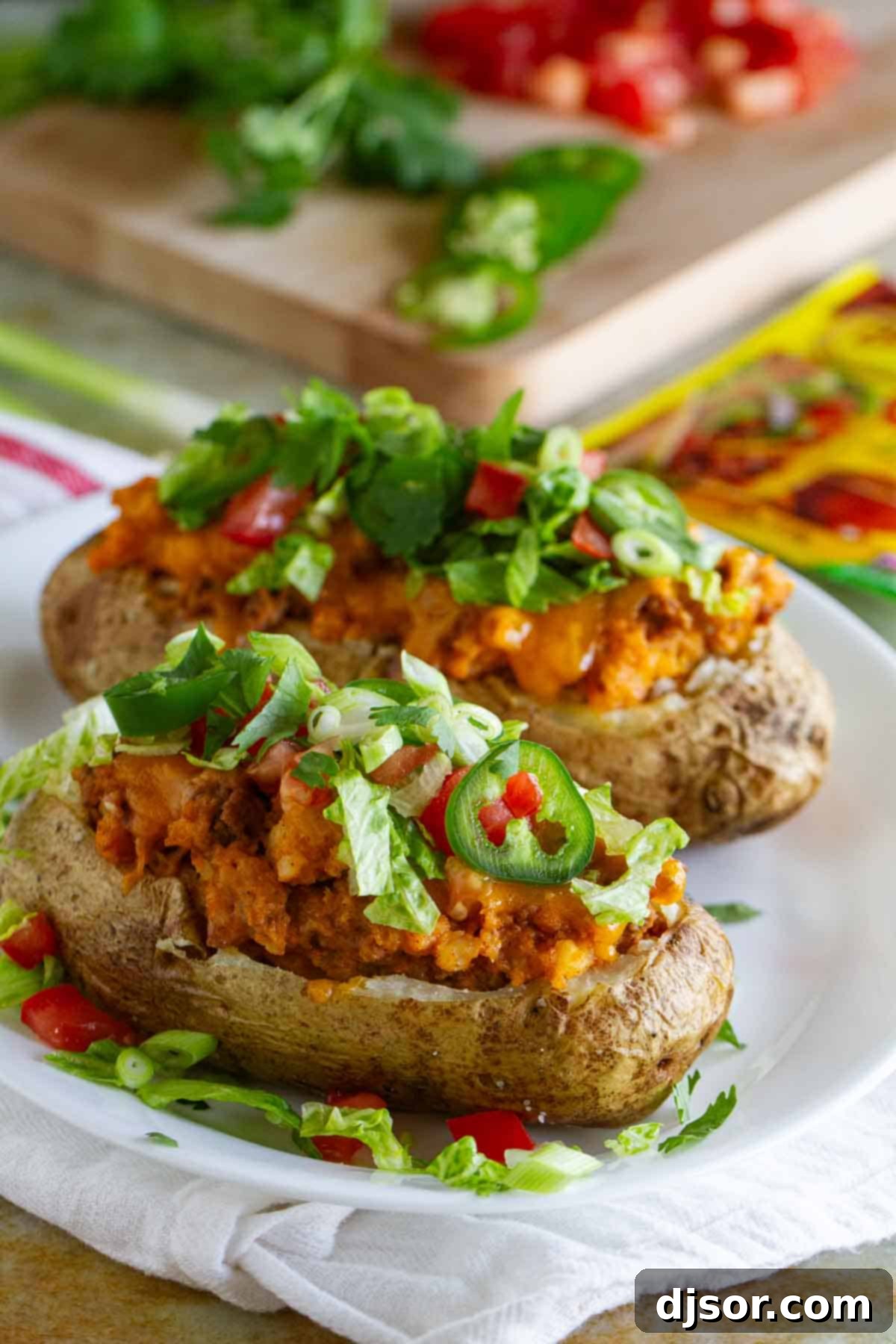 Two taco stuffed potatoes on a plate, topped with lettuce, tomatoes, and jalapeños.