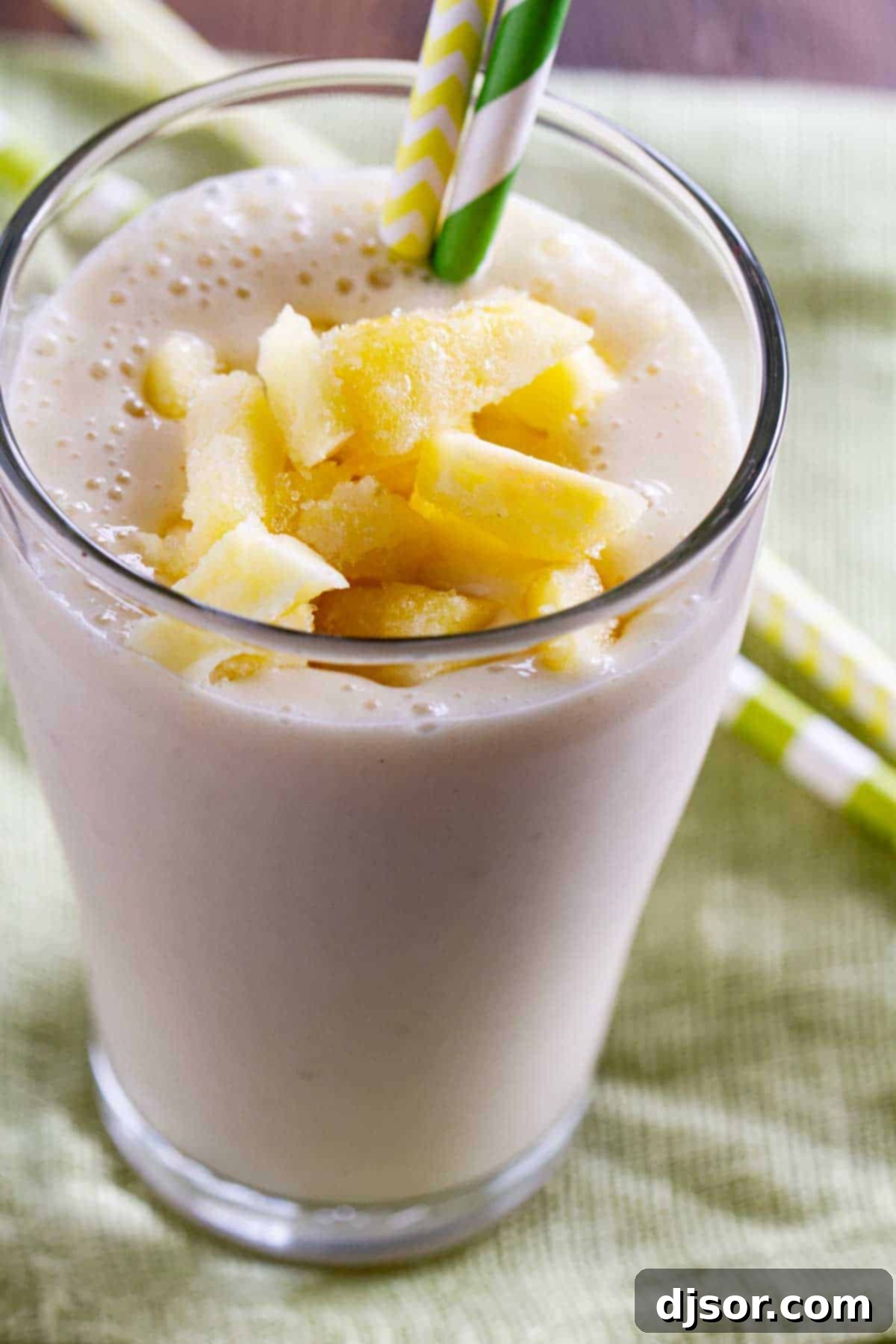 Glass filled with a vibrant yellow pineapple banana smoothie, garnished with fresh pineapple wedges and two straws, suggesting a shared, refreshing experience. A perfect healthy breakfast or tropical afternoon pick-me-up.