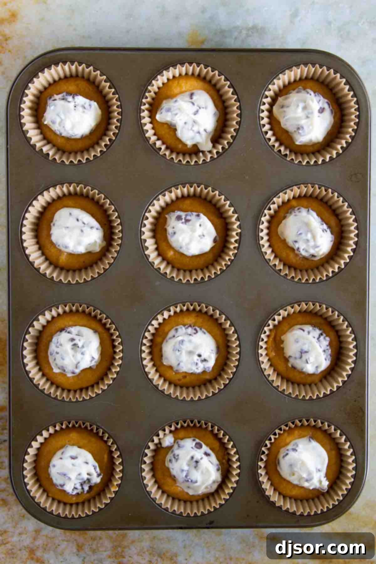 A muffin tin filled with layers of pumpkin batter and cheesecake filling, ready for baking into Pumpkin Cheesecake Spider Cupcakes.